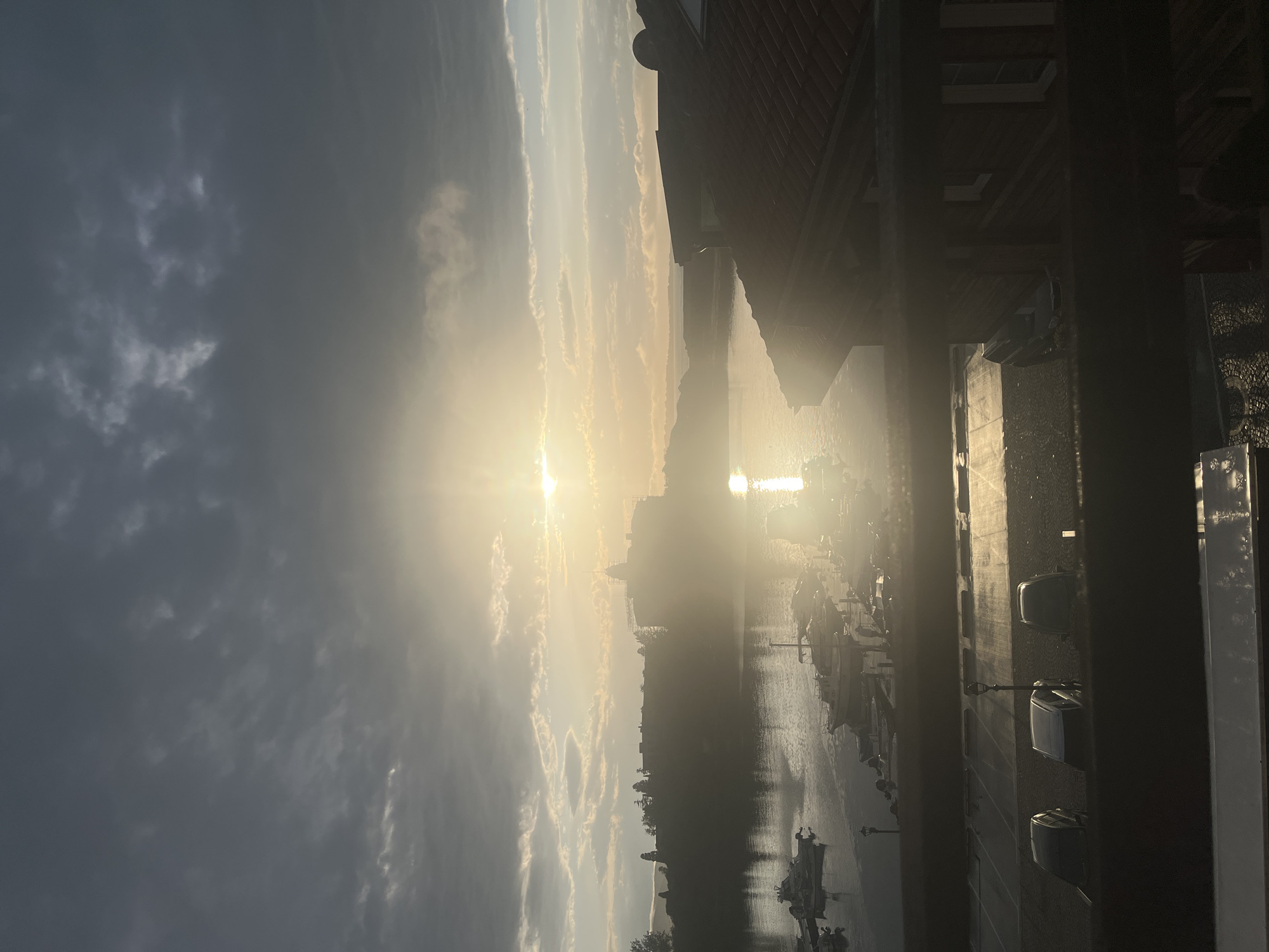 The image depicts a serene sunrise or sunset over a body of water, viewed from an elevated perspective. The scene is dominated by a very bright sun positioned towards the left side, casting a strong glare and illuminating the sky and water with a warm, golden glow.
The sky features a mix of clouds, ranging from darker, heavy clouds on the right to lighter, more ethereal wisps and streaks that are brightly lit by the sun, especially across the upper-left and central portions. The light reflects intensely off the water, creating a shimmering path directly beneath the sun.
On the water, numerous boats of varying sizes are visible, some appearing to be docked along a shore or marina on the right, while others are spread further across the expanse. In the mid-ground, a distant landmass or city skyline is silhouetted against the bright horizon, appearing as dark, indistinct shapes.
In the immediate foreground, dark, shadowed architectural elements, including a portion of a building with a reddish roof and vertical beams or pillars, frame the scene on the top and right, indicating the photograph was taken from within or adjacent to a structure. These foreground elements are heavily underexposed due to the intense backlight.
The time of day is clearly either early morning or late evening, given the low position of the sun and the quality of light. The weather appears partly cloudy with clear visibility. The atmosphere evokes a sense of peacefulness and natural beauty. No specific activity is clearly depicted, and no text is visible in the image.