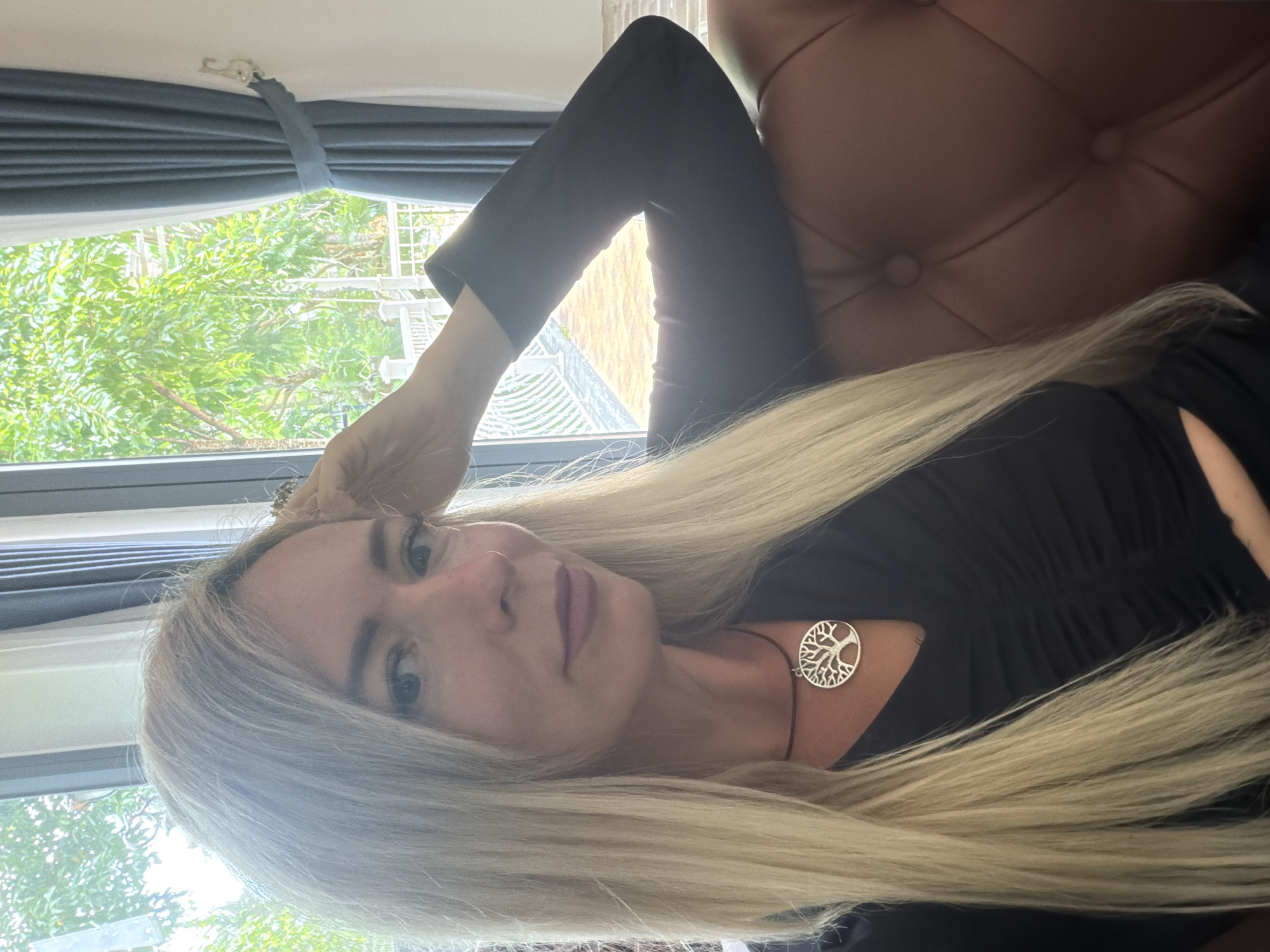 In a room with large windows overlooking lush greenery, a woman with long blonde hair and wearing a black dress is captured from the chest up. She is reclining on a tufted brown leather sofa, her right arm resting on the back of the sofa and her hand touching her head. She is wearing a necklace with a tree of life pendant. The room appears to be in a residential setting in Xuân Trường - Đà Lạt, Vietnam, evidenced by the tropical foliage visible through the windows and the style of the interior.