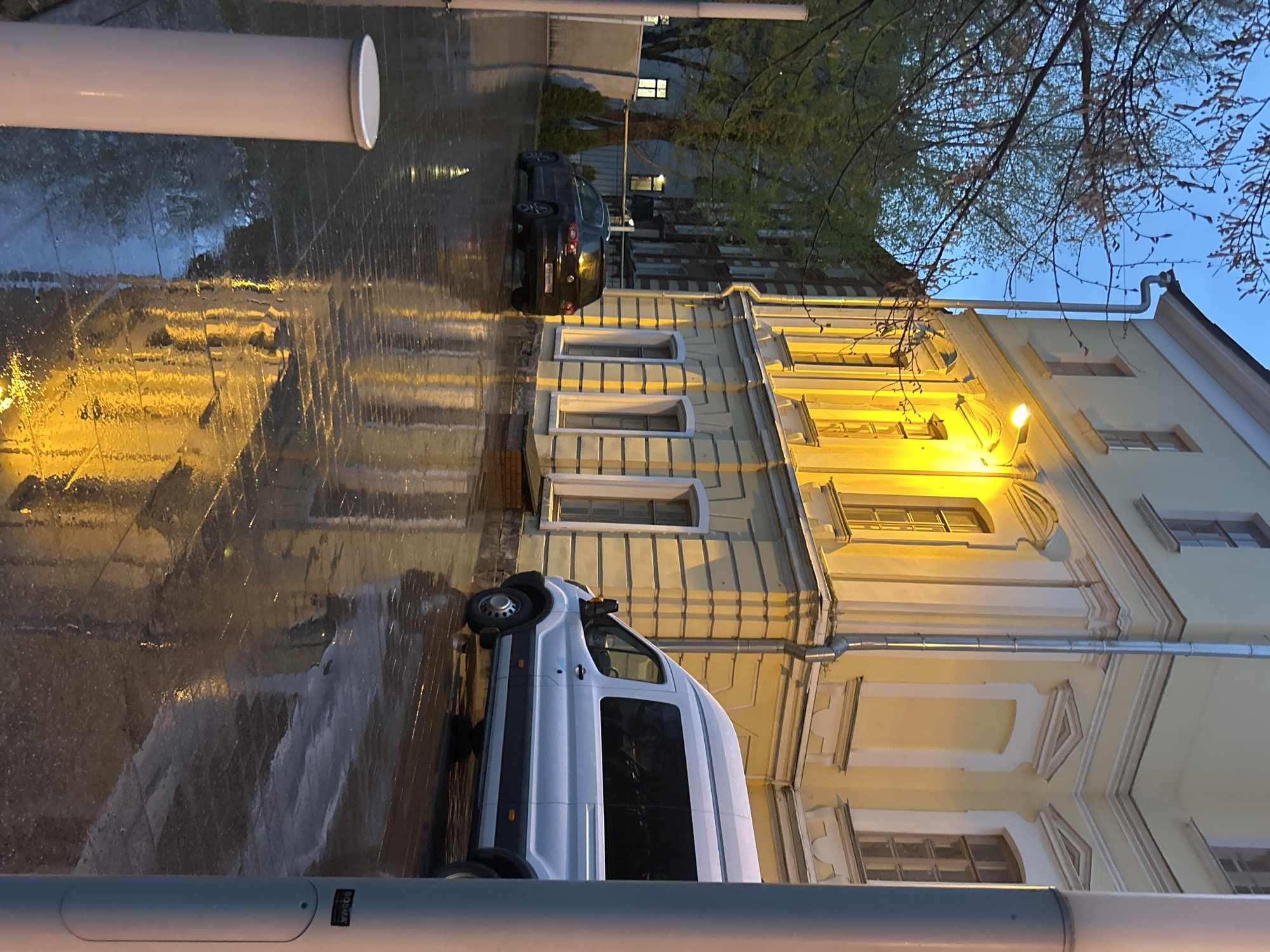 The image shows a street scene in Unknown, Unknown at dusk or dawn after a rain. A building with yellow walls and white trim is visible on the right. A white van is parked parallel to the building. Further down the street, a dark car is parked. The street is wet and reflects the yellow light from the building. The sky is a light blue color with bare tree branches visible in the upper right. In the foreground, part of a gray cylindrical structure is visible.