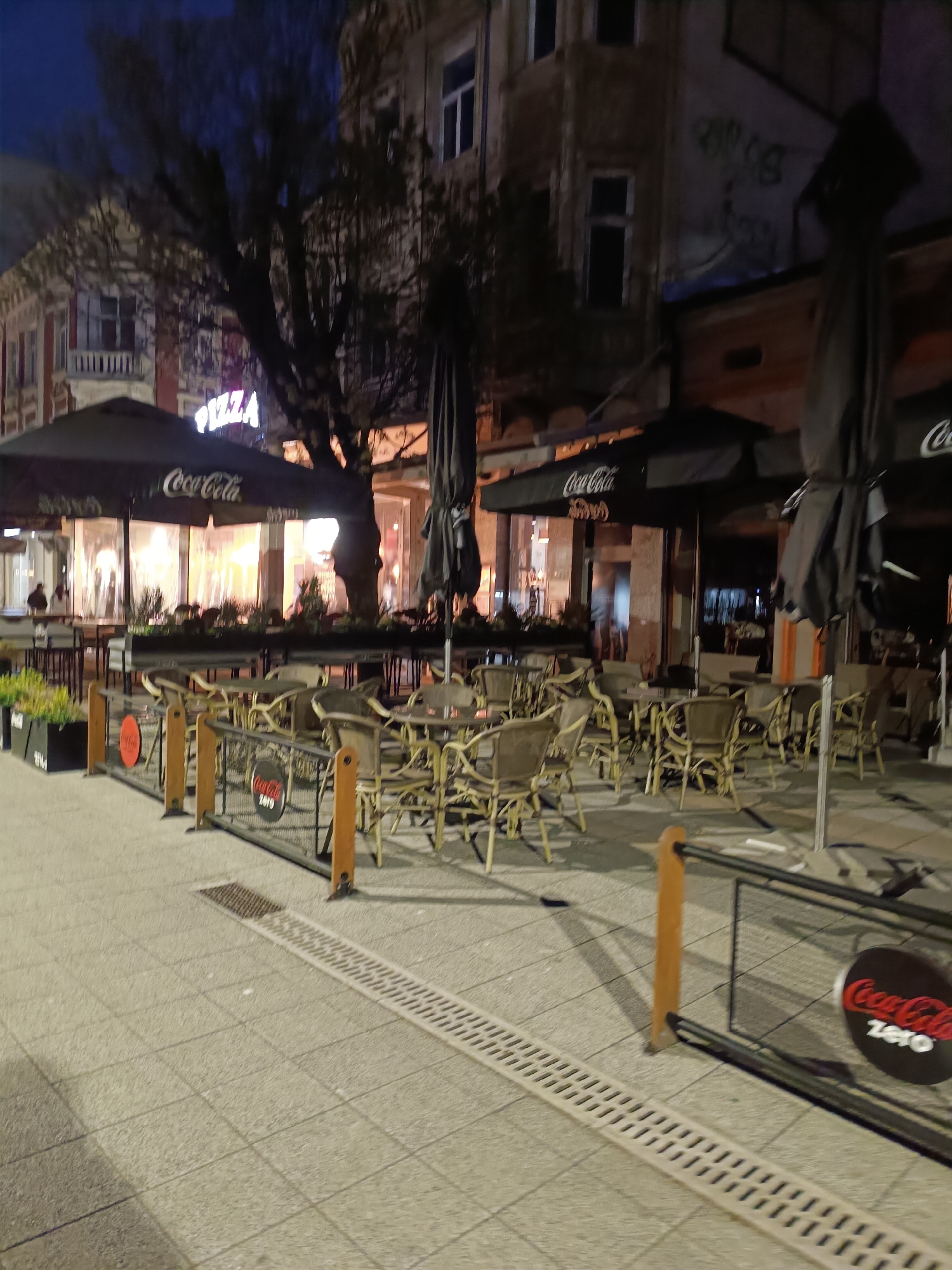 This image captures a quiet night scene of an outdoor dining area on a paved street in Pleven, Bulgaria. The time of day is clearly evening or night, indicated by the dark sky and artificial lighting from street lamps and the illuminated restaurant interior.
The central focus is an empty outdoor seating area, likely belonging to a restaurant or cafe. It is furnished with numerous light-colored chairs and tables, some neatly arranged, while others appear slightly askew. Several large, black outdoor umbrellas, some open and some folded, feature prominent "Coca-Cola" branding. Above one of these umbrellas, a glowing neon sign spells out "PIZZA," indicating the establishment's offerings.
The dining space is defined by a low, black metal fence with wooden posts, to which circular "Coca-Cola Zero" signs are attached. Planters with sparse greenery line parts of the perimeter. Behind the seating area, a large, bare tree stands with its branches silhouetted against the dark sky. The restaurant itself is housed in a multi-story building with illuminated windows, revealing a blurred figure inside. Another building to the right shows signs of age, including some faint graffiti on an upper wall. The street is paved with light-colored square tiles and features a long, gridded drainage channel.
Only a couple of indistinct, distant figures are barely visible in the background, reinforcing the quiet and deserted atmosphere of the late hour. The overall mood is one of stillness and anticipation, suggesting the area is either closed for the night or experiencing a significant lull in activity.