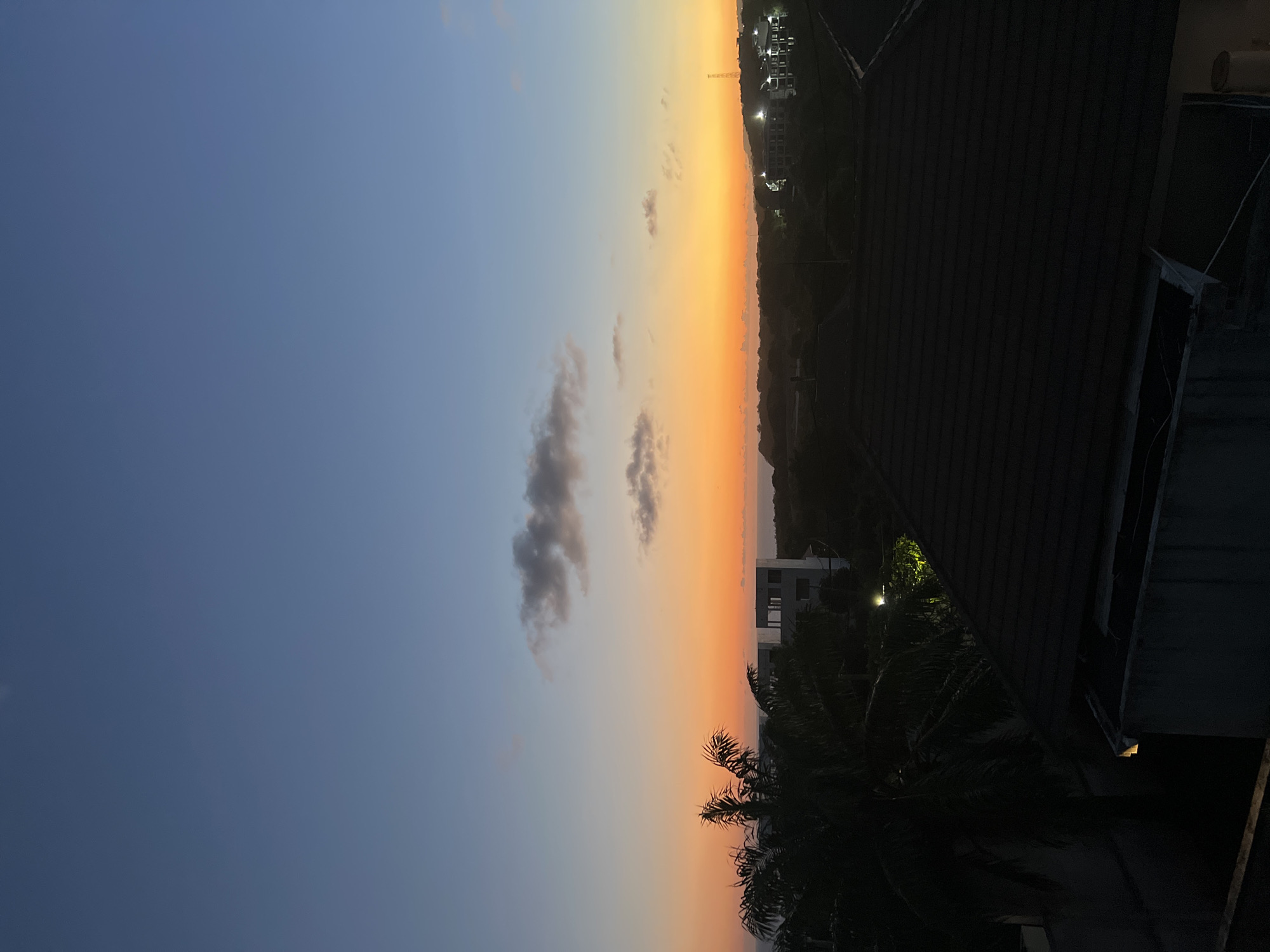 The image depicts a sunset over a landscape, rotated 90 degrees clockwise, showing a blend of natural beauty and urban elements. The dominant feature is the sky, which transitions from a deep blue at the top (east) to a vibrant orange and yellow hue towards the horizon (west), indicating the setting sun. A few small, dark, and wispy clouds are scattered across the orange-yellow portion of the sky.
In the foreground, the dark silhouette of a slanted roof structure occupies the bottom right, suggesting the vantage point is from a building or elevated position. Below this roof, dark foliage, likely palm trees, is visible, extending into the midground.
The horizon line is visible across the center, where the sky meets a distant, dark landmass, possibly hills or coastline. Several buildings are scattered across this landscape, some appearing as silhouettes, while others show illuminated windows and exterior lights, highlighting them against the darkening environment. A tall, crane-like structure is faintly visible in the distance near some illuminated buildings. Another, more rectangular building is closer to the horizon on the left side of the frame.
The scene conveys a peaceful twilight atmosphere. There are no people visible, nor any discernible activity or visible text. The presence of palm trees and the general architecture hint at a tropical or subtropical location.