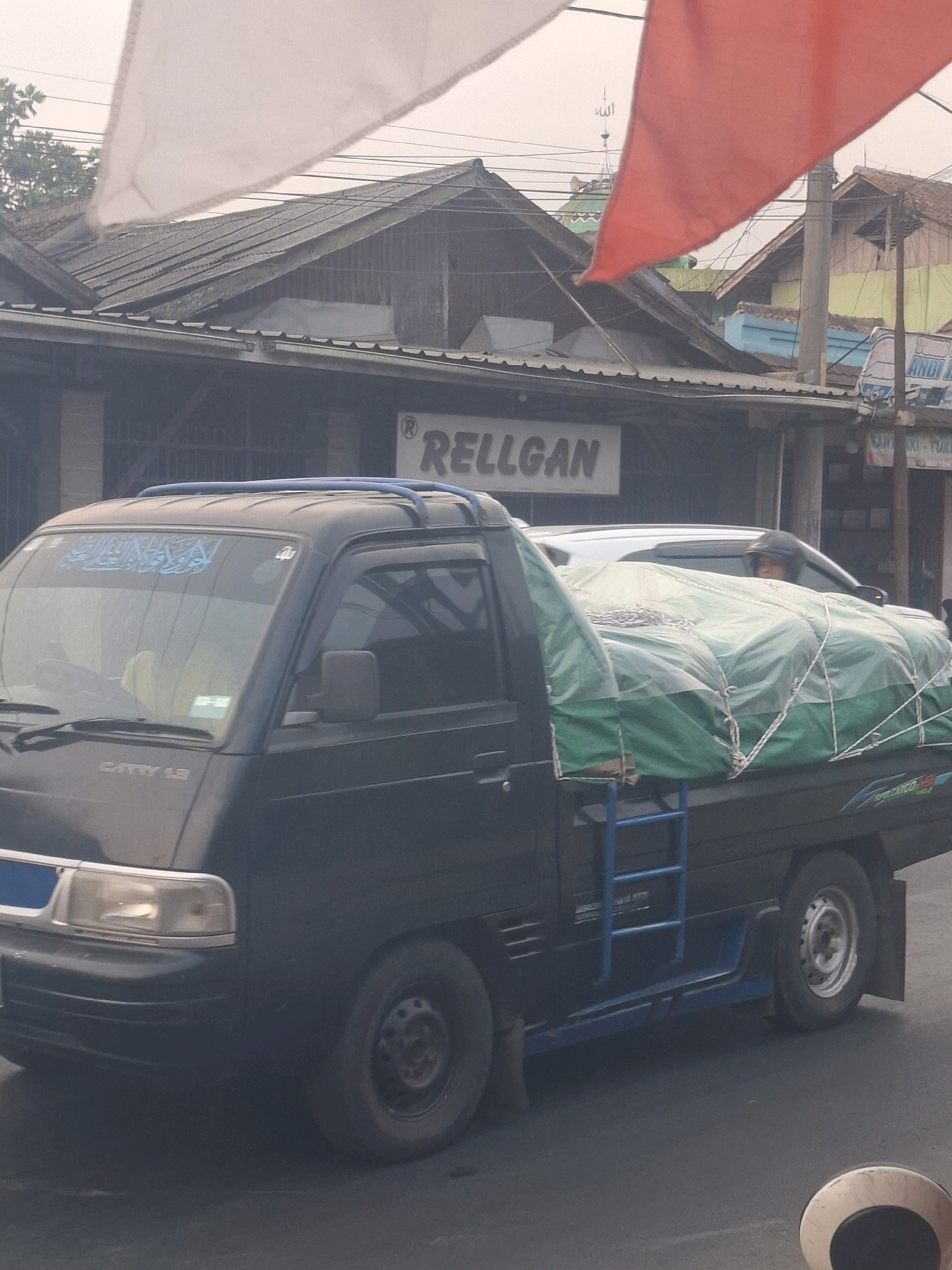 In the city of Cijalingan, Indonesia, a dark blue pick-up truck with a covered cargo bed is parked on a street. The cargo is draped in a green tarp secured by white ropes. A blue metal ladder is attached to the side of the truck. The truck's windshield has blue text written on it, and the words "CARRY 1.3" are visible on the hood. In the background, buildings with tiled roofs are visible, along with utility poles and wires. A red and white flag are visible in the upper portion of the image, and a mosque dome can be seen behind the buildings. The sky appears overcast.
