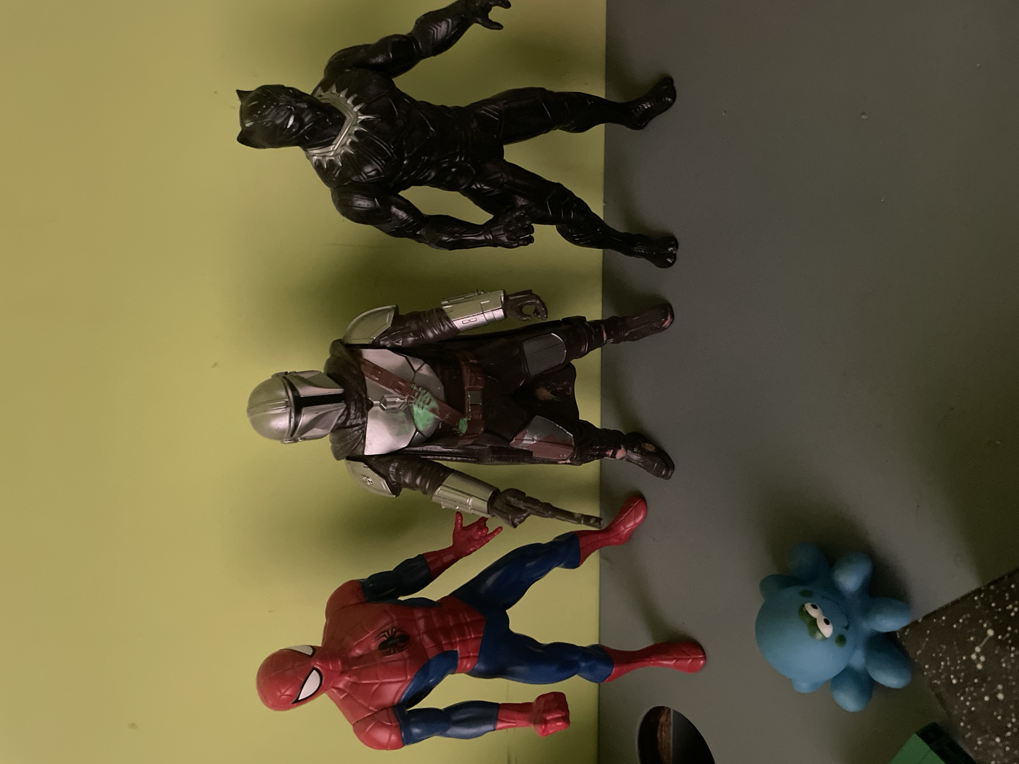 The image shows four figurines on a two-toned surface, likely a table or shelf. The upper portion of the surface is a pale green color, and the lower portion is gray. At the top is a black panther figurine in a leaping pose. Below it is a Mandalorian figurine lying horizontally with a blaster rifle. Next is a Spider-Man figurine posed as if crawling. To the right of Spider-Man, there is a small, blue octopus-like figurine. The location is not specified.