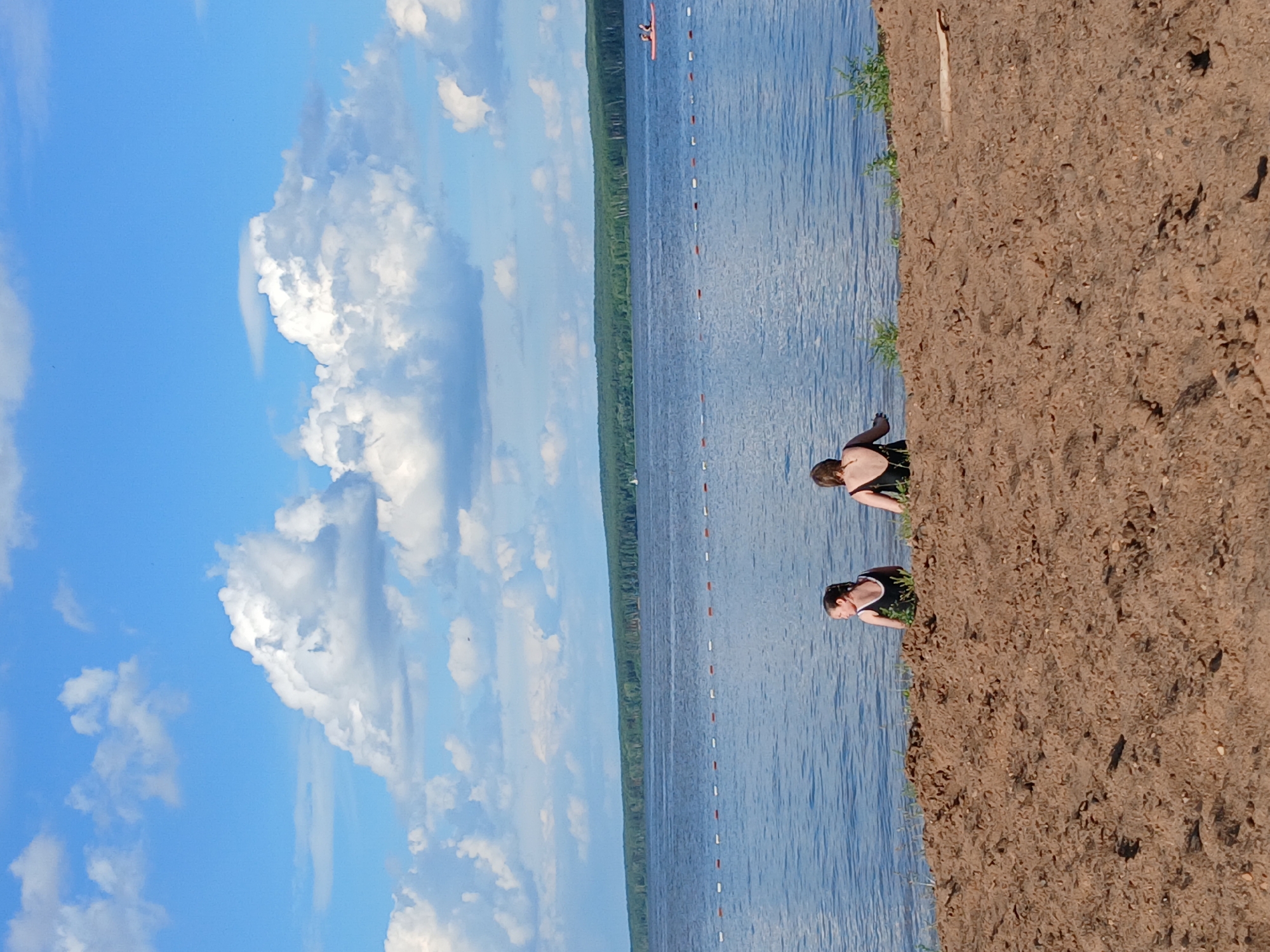 The image depicts a scenic view of a calm, blue lake on a sunny day. The water extends towards a tree-lined horizon, with a few white, puffy clouds dotting the vast expanse of the sky. In the foreground, a sandy beach is visible, with a few individuals relaxing near the water's edge.
Two people are seen on the beach, appearing to be enjoying the warm weather. One person is partially visible, wearing a dark-colored swimsuit and looking out at the water. Another person, further down the beach and closer to the water, is also wearing a dark swimsuit and is crouched down, possibly preparing to enter the water or observing something in the distance. The sandy texture of the beach is detailed, with scattered small stones and patches of green grass growing along the shoreline.
The water is calm with a slight ripple, reflecting the bright blue of the sky. A line of buoys floats on the surface of the lake, marking a designated swimming area. In the distance, a small orange kayak with one person paddling can be seen on the water, adding a touch of activity to the otherwise tranquil scene. The overall mood of the image is peaceful and relaxing, suggesting a pleasant summer day spent by the lake. The presence of greenery on the far shore and the clear blue sky indicate fair weather, likely mid-day given the brightness of the sunlight. The location is identified as Chaikovsky, Russia. There is no visible text in the image.
