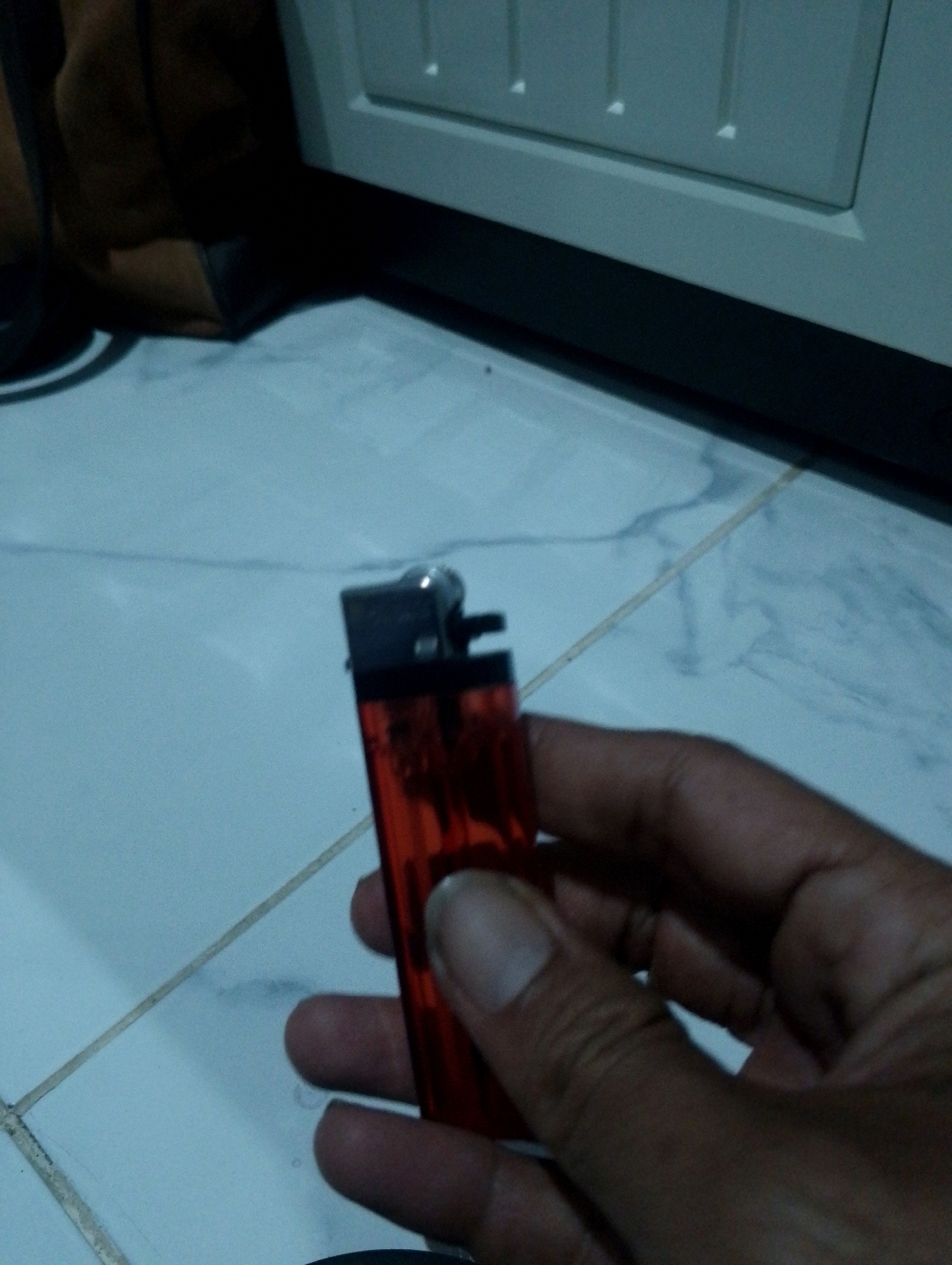 A close-up shot shows a hand holding a red cigarette lighter against a tiled floor. The lighter is partially transparent, revealing the fuel reservoir. The tiled floor appears to be a light blue or gray with darker grout lines. In the background, a portion of a cream-colored cabinet or furniture piece is visible. The lighting is dim, and the overall image has a grainy quality. The location is specified as Sumbergirang, Indonesia.