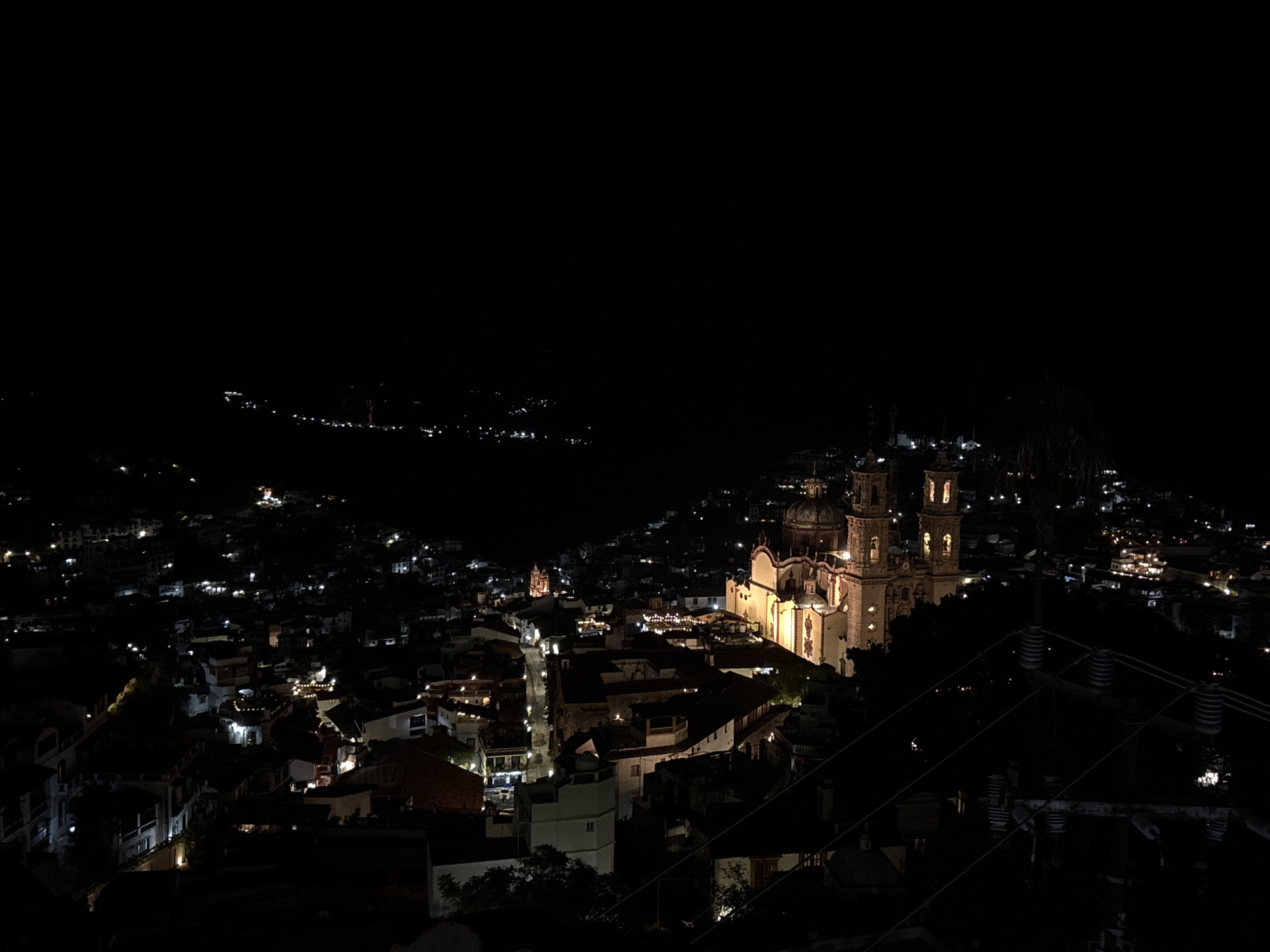 The image shows a nighttime aerial view of Taxco, Mexico. The prominent subject is the illuminated Parroquia de Santa Prisca y San Sebastián, a baroque church with twin towers and a large dome, glowing warmly against the dark sky. The surrounding city is visible as a collection of lights scattered across hillsides, indicating densely packed buildings. Streets are illuminated by streetlights and lights emanating from windows. The foreground features dark silhouettes of trees and power lines, suggesting the vantage point is from a higher elevation. The overall scene depicts a peaceful, glowing urban landscape at night.