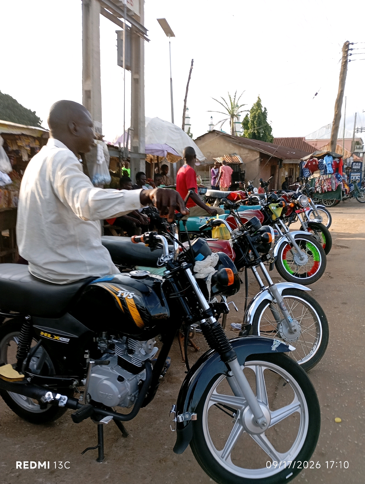 This is a medium shot, eye-level, outdoor, daytime photograph taken in Shendam, Nigeria. The scene depicts a row of motorcycles parked on a dirt road. In the foreground, a Black man wearing a light-colored long-sleeved shirt is seated on a black motorcycle, with his left hand on the handlebars. The motorcycle has "TVS" branding and an orange and black design on its fuel tank. Behind this motorcycle, several other motorcycles are visible, parked in a line. Some of these motorcycles are colorful, with red, green, and white accents on their wheels. In the background, there are buildings with corrugated metal roofs, market stalls displaying clothing, and people milling about. The sky is overcast. A wooden utility pole is visible on the right side of the image. The lower left corner of the image shows "REDMI 13C" and the lower right corner shows "09/17/2026 17:10".