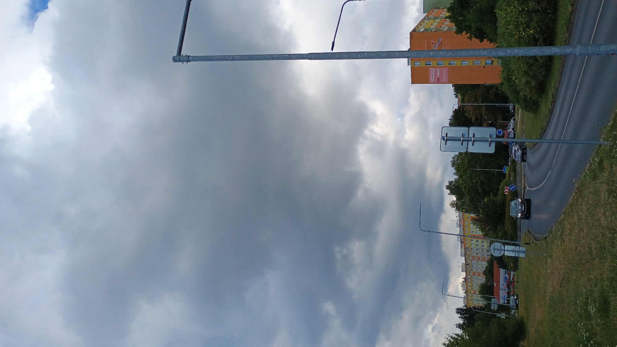 The image depicts a street scene in Sokolov, Czechia, under a cloudy sky. A multi-lane road curves through the frame, with grass and trees bordering the left side. Several cars are visible on the road, including a dark SUV in the foreground and a lighter car in the background. Streetlights and traffic signs are also present. In the background, apartment buildings with colorful facades stand among trees. The overall impression is of a residential area within a city, with traffic and urban infrastructure.