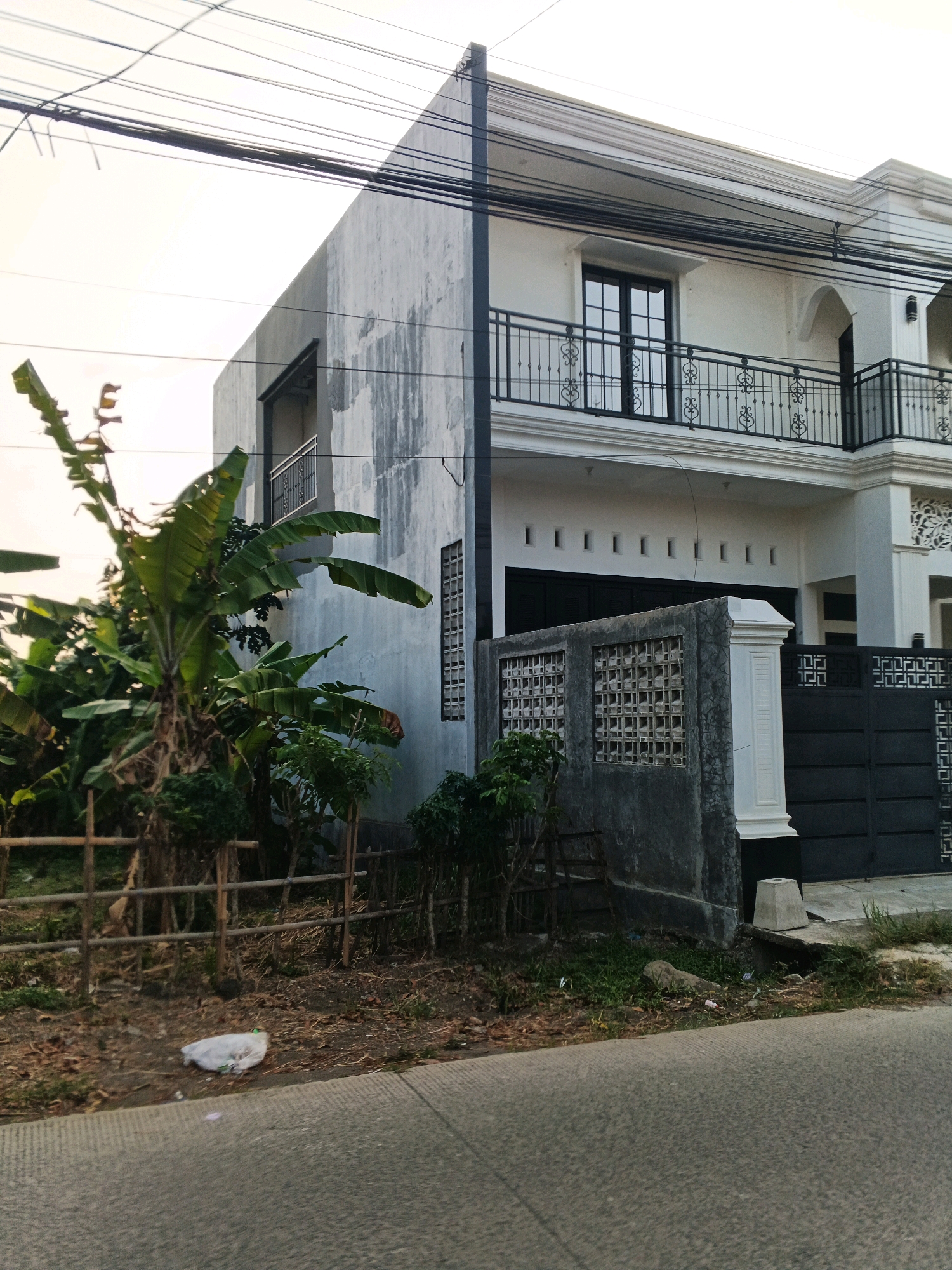 The image depicts a two-story residential building with a modern facade, located on a street in Planjan, Indonesia. The building is painted white, with dark-framed windows and decorative black metal railings on the balconies. A large black metal gate is visible at the front, partially obscured by a concrete fence with square openings. Lush green banana trees and other vegetation are situated in front of the building, separated from the street by a rustic wooden fence. The foreground shows a paved road with a white plastic bag lying on the side. Overhead power lines crisscross the sky, and the overall lighting suggests it is either early morning or late afternoon. The scene appears to be a residential area, with no people or vehicles actively present in the frame.