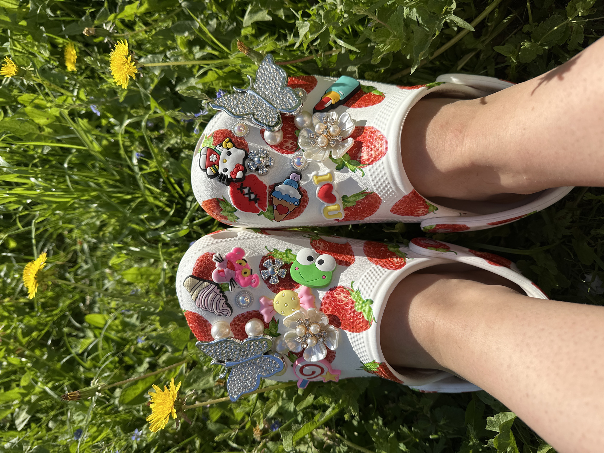 A close-up, slightly high-angle shot shows two feet clad in white Crocs-style shoes decorated with charms, set against a background of green grass interspersed with yellow dandelions. The shoes are patterned with red strawberries and adorned with a variety of charms, including a glittering butterfly, a Hello Kitty figure, a frog, a flower, a piece of candy, and other decorative elements. The wearer's legs are fair-skinned and visible from the ankles down, partly shadowed. The setting appears to be an outdoor grassy area, potentially a yard or park, in bright daylight. The exact location in Nizhny Novgorod, Russia, cannot be determined from the given visual information.