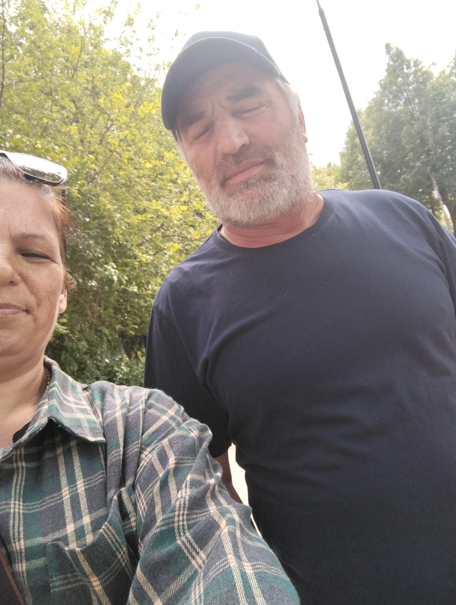 The image shows two people, a woman on the left and a man on the right, standing outdoors. The woman is wearing a green and blue plaid shirt and sunglasses on her head. The man is wearing a navy blue t-shirt and a black baseball cap. They are both looking towards the camera, and the man's eyes are closed. The background is out of focus, but appears to be a park or wooded area with green trees and a bright sky. The image was taken in Kingisepp, Russia.