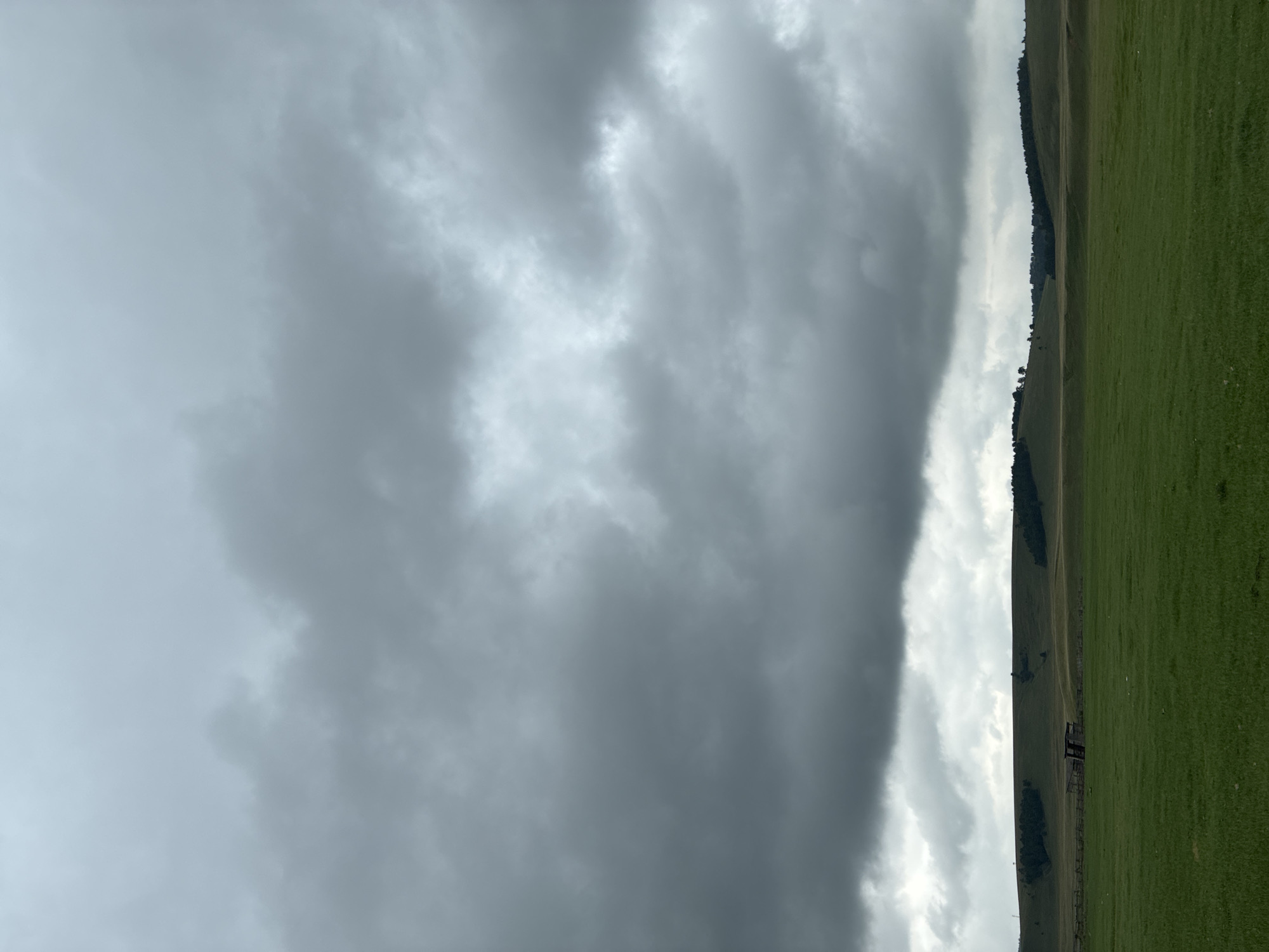 The image depicts a wide, grassy field under a heavily overcast sky. The foreground is dominated by vibrant green grass. In the middle ground, a low, rolling hill is visible, dotted with scattered trees and a small, derelict wooden structure resembling a shed or a small barn. The horizon is marked by the undulating line of the hill. The sky is filled with dark, heavy gray clouds, suggesting an imminent rain or a generally gloomy weather condition. The scene appears to be located in an open, rural area.