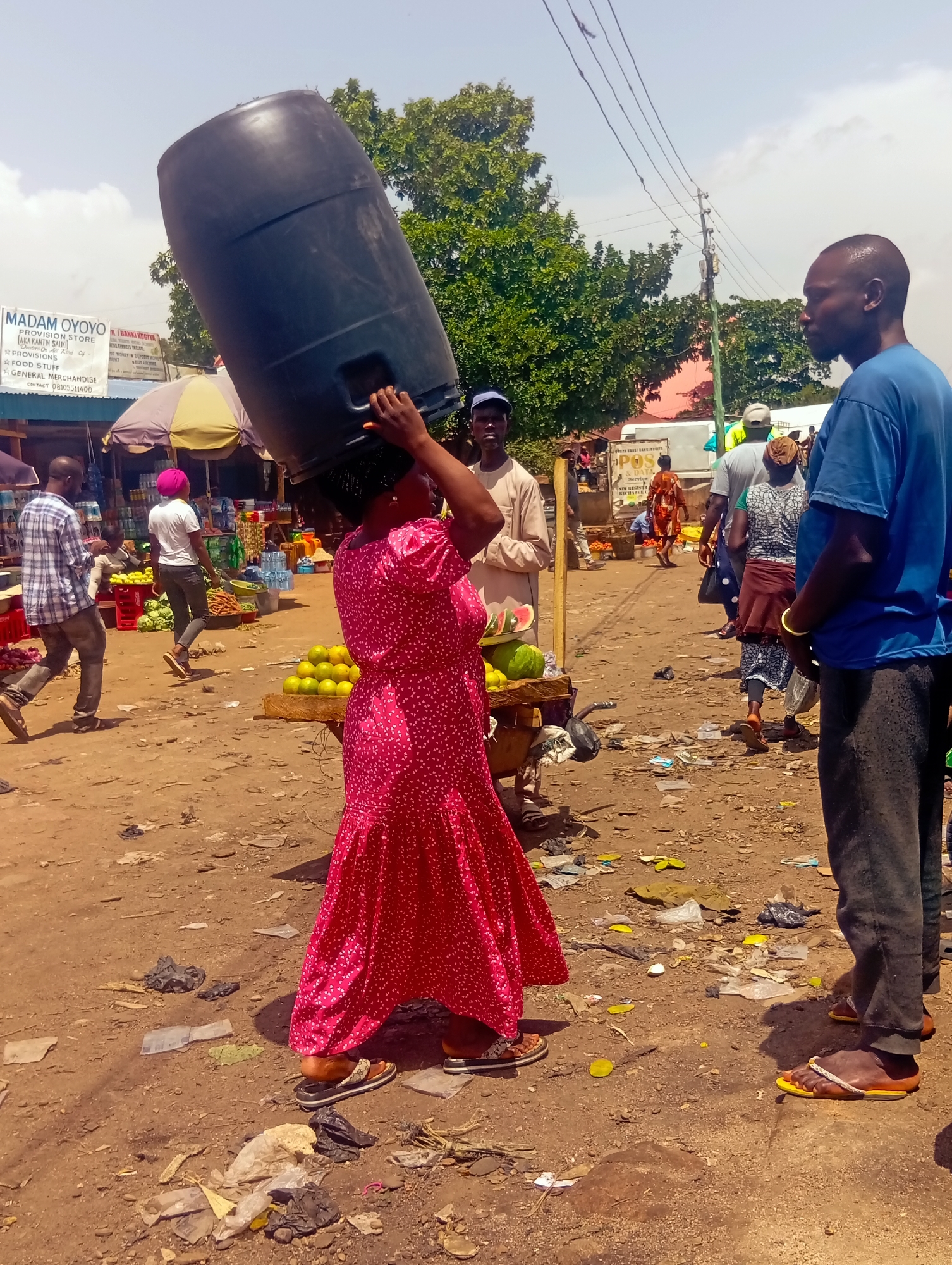 In the city of Bukuru, Nigeria, a woman in a red patterned dress carries a large black plastic barrel on her head. She is walking towards the right of the frame, appearing to be in a bustling outdoor market. To her left, a man in a blue shirt and dark pants stands looking at her, with his right hand on his hip. Behind him, a man in a plaid shirt walks away from the camera. In the background, other people can be seen navigating the market, with stalls selling produce like oranges and watermelons visible. The ground is dusty and littered with debris, suggesting a well-worn path in a busy commercial area. The sky above is partly cloudy. A sign for "MADAM OYOYO PROVISION STORE" is visible in the background, indicating the presence of businesses in the vicinity.