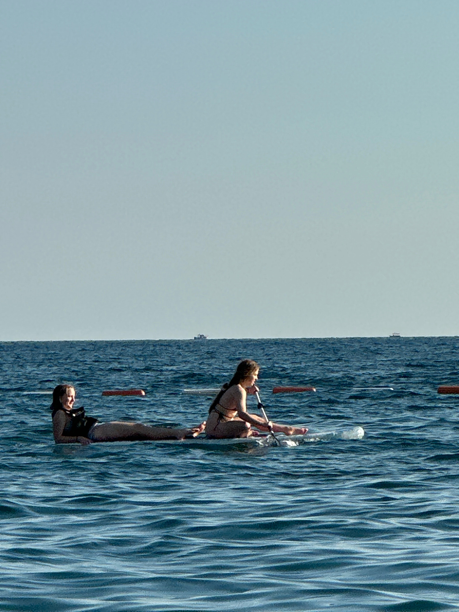 Two young women are on a transparent paddleboard in the ocean. The woman on the left is lying down on her stomach and looking to her left with a smile. The woman on the right is sitting up and holding a paddle, looking towards the left. The paddleboard is floating on dark blue water with small waves. In the background, there are several orange buoys and a few distant boats on the horizon. The sky is clear and light blue. The location is the coast of Boreti, Montenegro.
