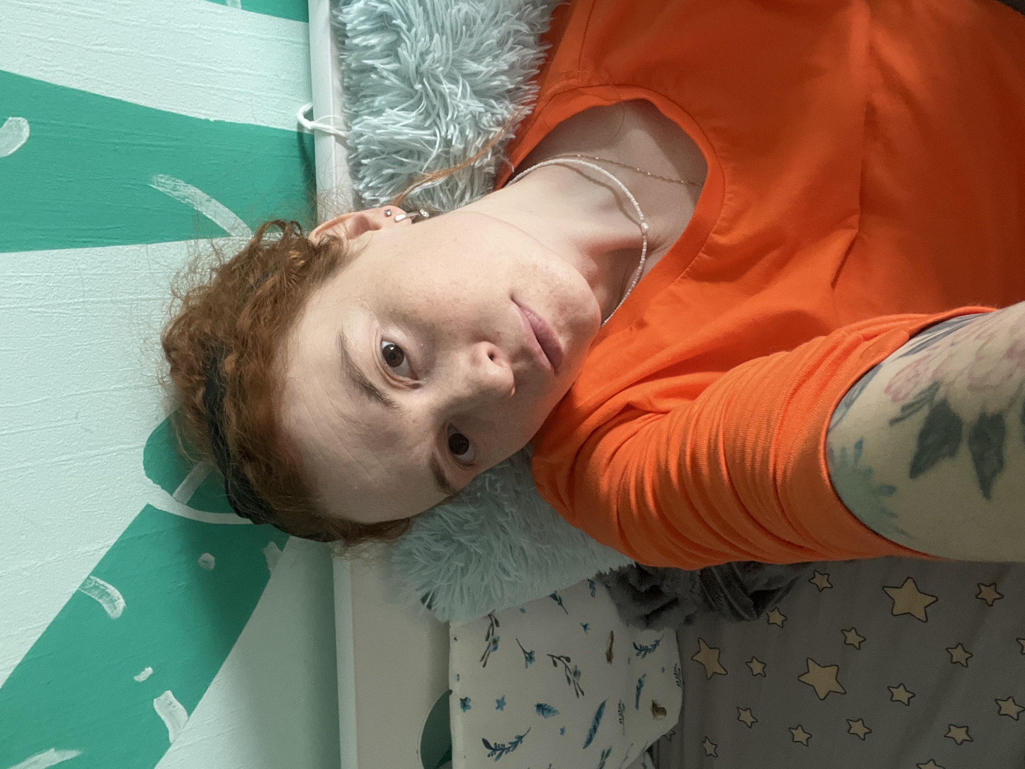 A woman with fair skin, auburn hair, and wearing an orange long-sleeved shirt is lying on her side, likely on a bed or similar surface. She is looking directly at the camera. Her right arm is visible, showing a tattoo on her upper arm. She is wearing necklaces. Her head is resting on a light blue, fluffy pillow. The background features a wall painted with green and white stripes. The bed linens include a light grey fabric with yellow stars and a white fabric with blue floral and feather patterns. The location is likely an interior space, possibly in a residence. The city is Chaikovsky, Russia.