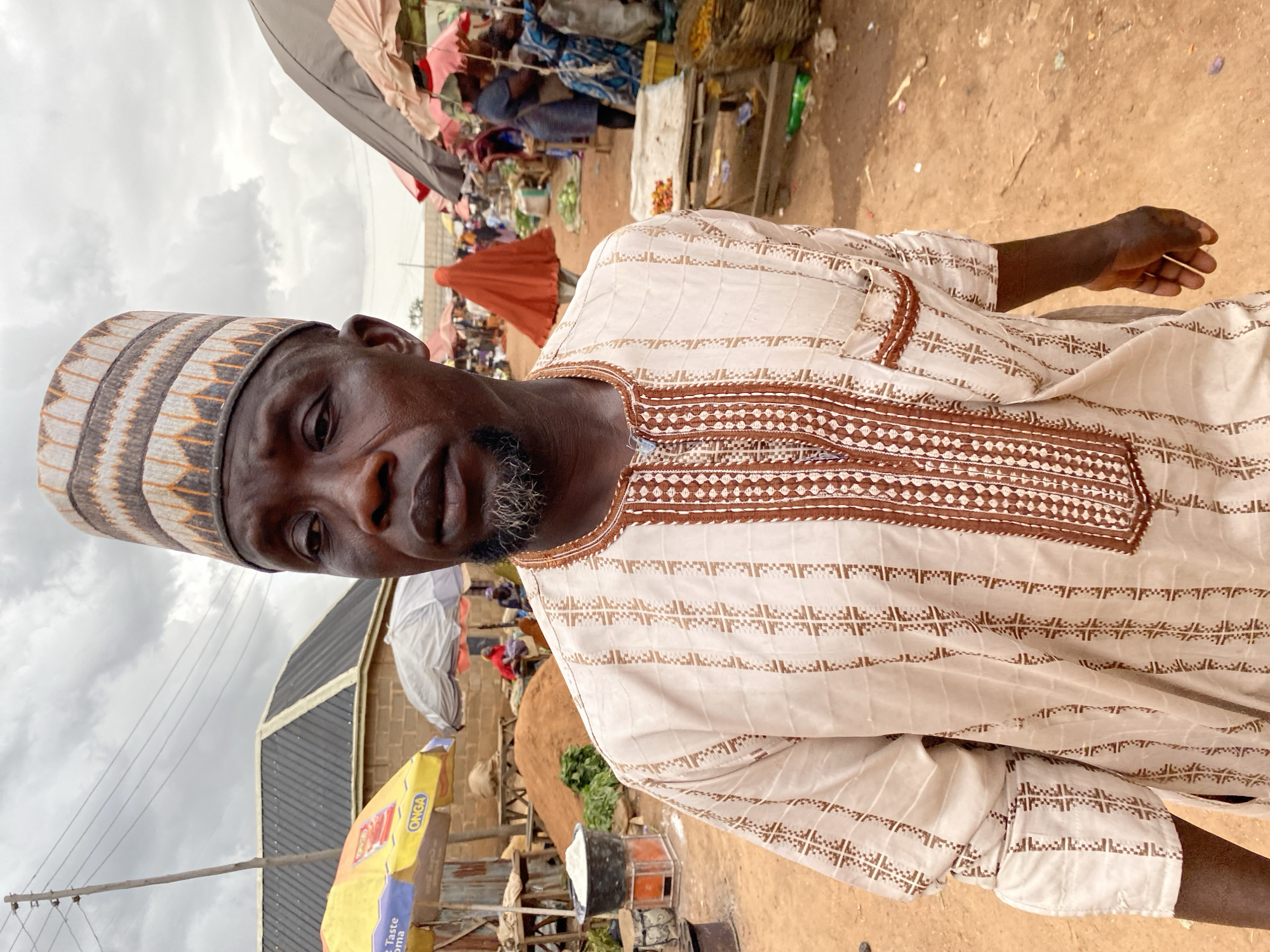 In Bukuru, Nigeria, a man stands in a market under a cloudy sky. He is wearing a light-colored robe with brown trim and a patterned cap. The market is bustling with people and stalls, indicated by the presence of umbrellas, baskets of goods, and mounds of dirt. The ground is made of red earth, and various structures, including a brick building and a corrugated metal roof, are visible in the background.
