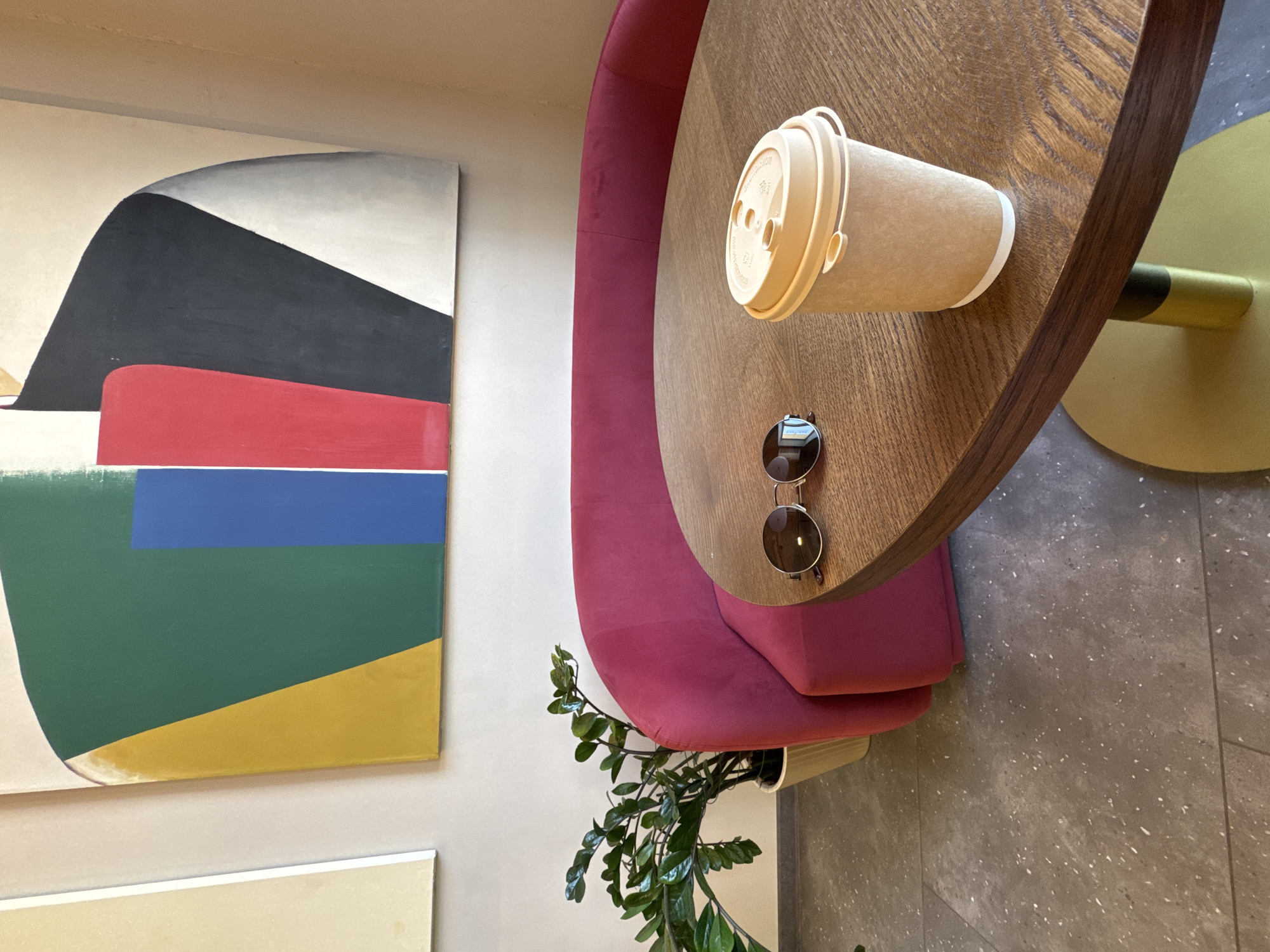 The image depicts an interior scene, possibly a cafe or a waiting area, with modern abstract art on the wall. In the foreground, a round wooden table is set with a disposable coffee cup and a pair of sunglasses. Behind the table, a vibrant pink upholstered sofa curves around, partially visible. To the left of the sofa, a potted green plant with thick, waxy leaves stands on a speckled gray floor. The overall ambiance is contemporary and stylish, with a focus on bold colors and clean lines. The location is specified as Krasnodar, Russia. There are no people present in the image.