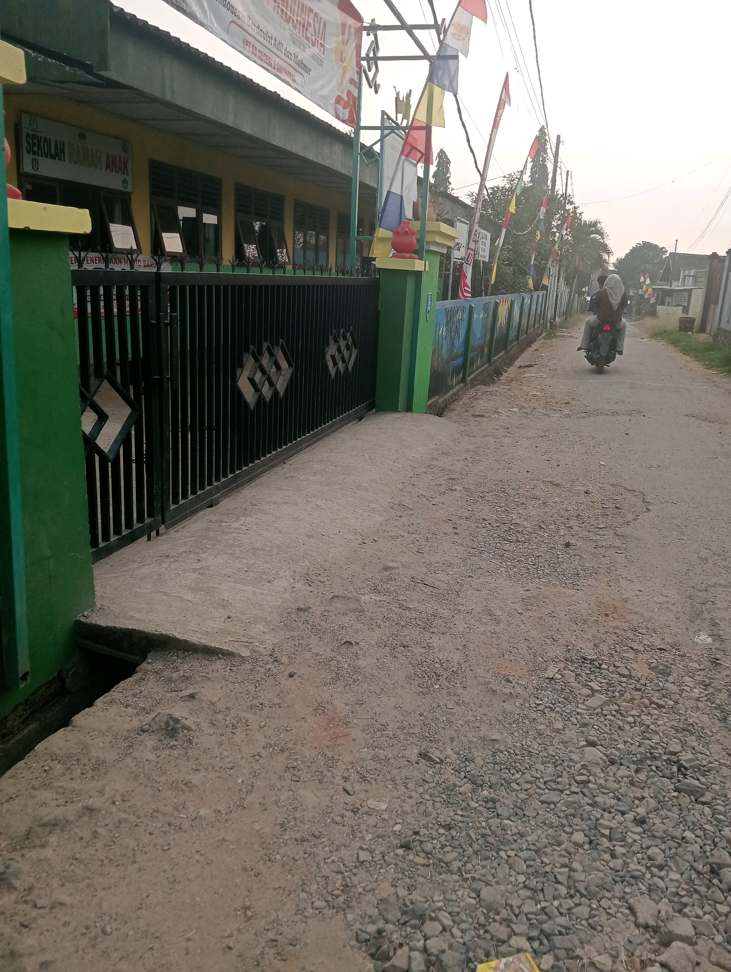 The image depicts the exterior of a school building in Pringsewu, Indonesia. The school appears to be a "Sekolah Ramah Anak" (Child-Friendly School). In the foreground, a green-painted gate with a black metal fence is visible, leading to the school grounds. The fence has decorative diamond-shaped patterns. The ground in front of the gate is a rough, unpaved surface composed of dirt and small stones.
Beyond the gate, the school building has a yellow facade with several windows. A banner above the building reads "INDONESIA" and mentions "UPT SD NEGERI 2 SIDOMULYO," indicating this is public elementary school number 2 in Sidomulyo.
To the right of the school, a narrow, unpaved road stretches into the distance. Two individuals are riding a motorcycle on this road, moving away from the viewer. They are both wearing headscarves. Along the side of the road, a mural-painted wall borders the school property. Various flags, including what appear to be Indonesian flags and other colorful banners, are strung along poles and wires above the school and road. The overall environment suggests an outdoor scene in a semi-urban or rural area, with a slightly overcast sky.