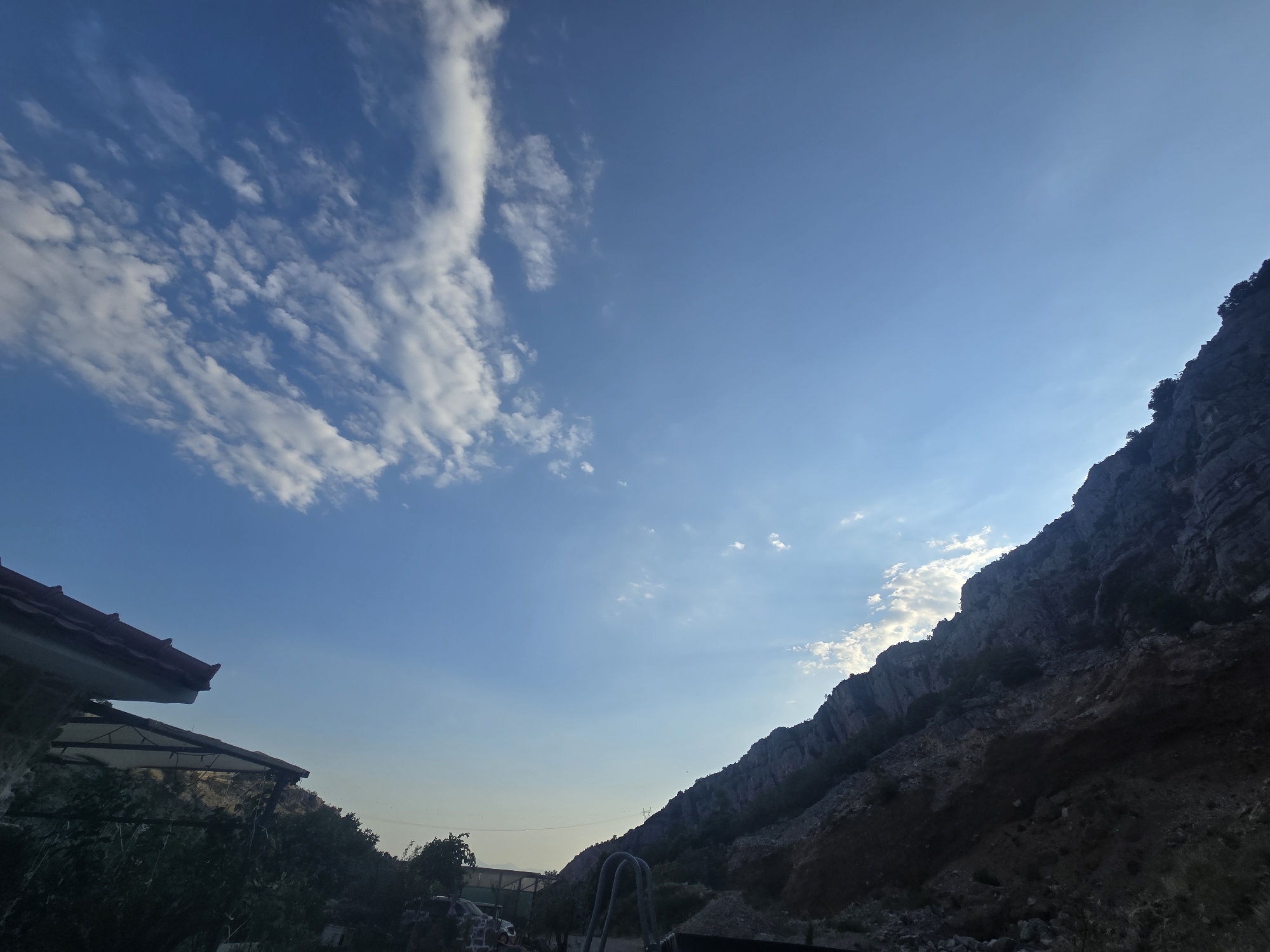 The image depicts a clear blue sky with scattered white clouds, casting subtle shadows on a rugged, rocky mountainside that dominates the right side of the frame. In the foreground, a building with a red-tiled roof and an overhang is visible on the left, partially obscured by dark green foliage. The mountainous terrain appears arid, with sparse vegetation clinging to its slopes. The overall lighting suggests it is either early morning or late afternoon. The scene evokes a natural, somewhat undeveloped environment, likely on the outskirts of a settlement given the presence of the building. This location could be near the Zeta valley or the outskirts of Podgorica, Montenegro, given the characteristic karst topography and vegetation. There are no people or animals visible in the image.