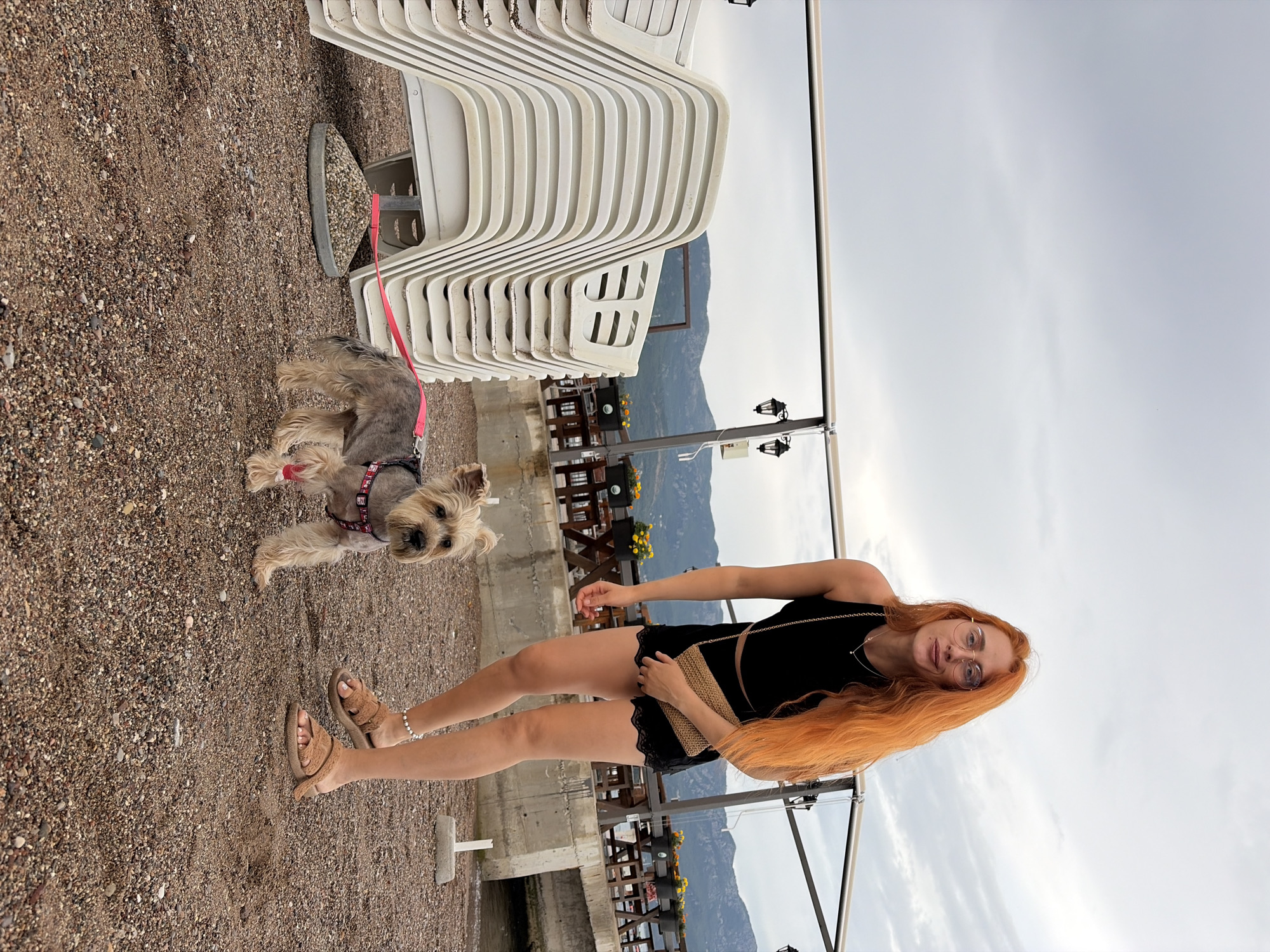 A young woman with long, bright orange hair and glasses is standing on a gravelly surface, possibly a beach or promenade, holding a small dog on a pink leash. She is wearing a black tank top, black shorts with lace trim, and brown furry slides. She has a woven clutch bag over her shoulder. The dog is a small terrier mix with a grey and tan coat, wearing a patterned harness.
Behind the woman and dog, a stack of white plastic lounge chairs is visible, suggesting a beach resort or outdoor seating area. In the background, there are wooden tables and chairs, indicative of an outdoor restaurant or cafe. Beyond that, a mountain range can be seen under an overcast sky. Two black lampposts are also present, one with a small electrical box attached.
The time of day appears to be late afternoon or early evening, given the soft light and cloudy conditions. The overall mood is relaxed, as the woman and dog are enjoying a casual outing. The location cues, such as the gravelly ground, lounge chairs, and mountainous backdrop, are consistent with a coastal area in Budva, Montenegro. There is no visible text in the image.
