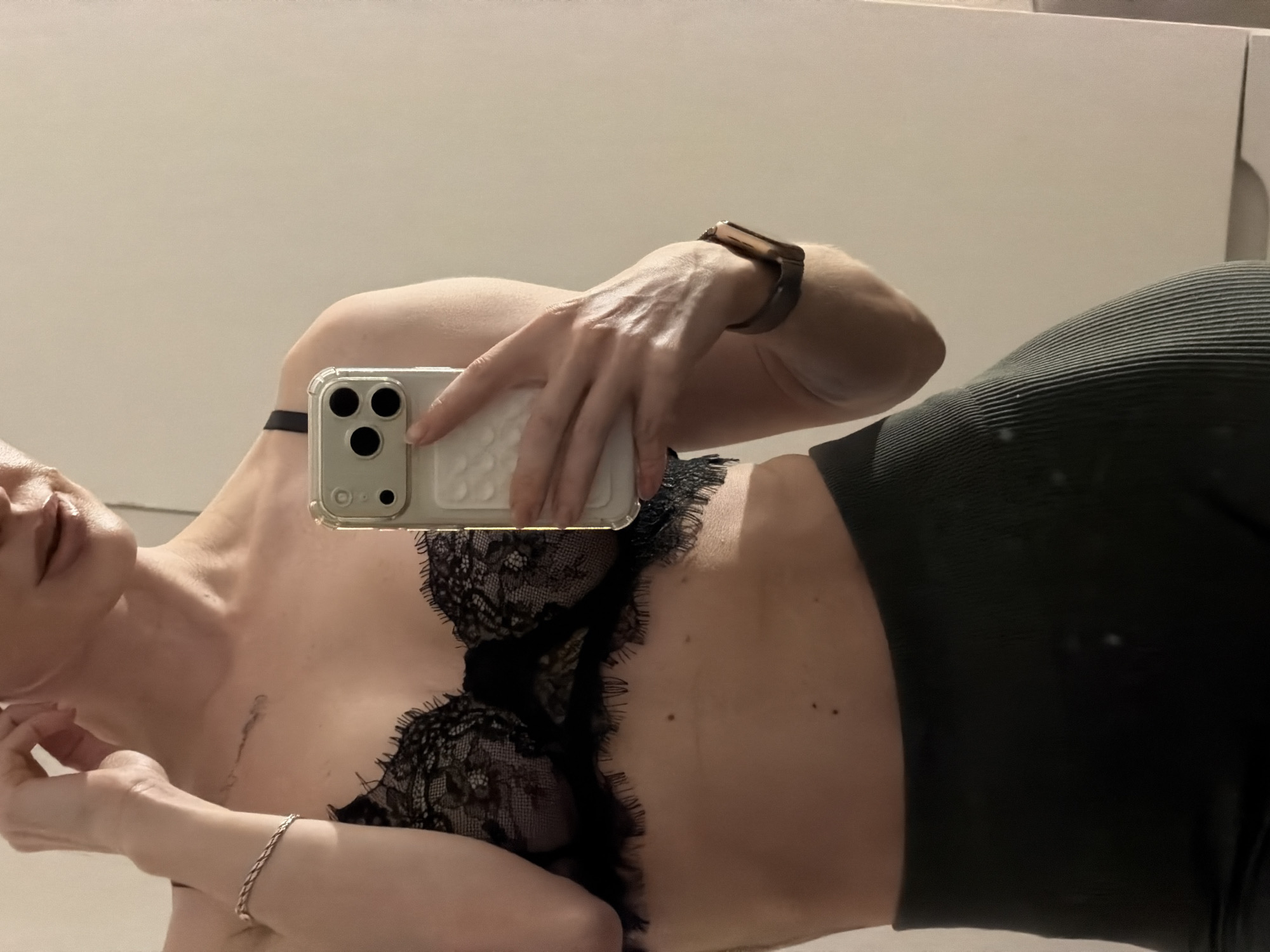 A person, likely female, is shown taking a mirror selfie while lying on their side. They are wearing a black lace bra and dark, ribbed leggings or pants. In their right hand, they hold a light-colored smartphone with a clear case, featuring three camera lenses. A smartwatch with a metallic-toned case and band is visible on their right wrist, and a silver-colored braided bracelet adorns their left wrist.
Notable physical details include fair skin, several moles on the torso, and a distinct scar or mark on the upper chest/collarbone area. The person's lips are slightly parted. The background consists of a plain, light-colored wall or surface, indicating an indoor setting. There is no visible text in the image. The time of day and specific emotions are not discernible from the photograph.