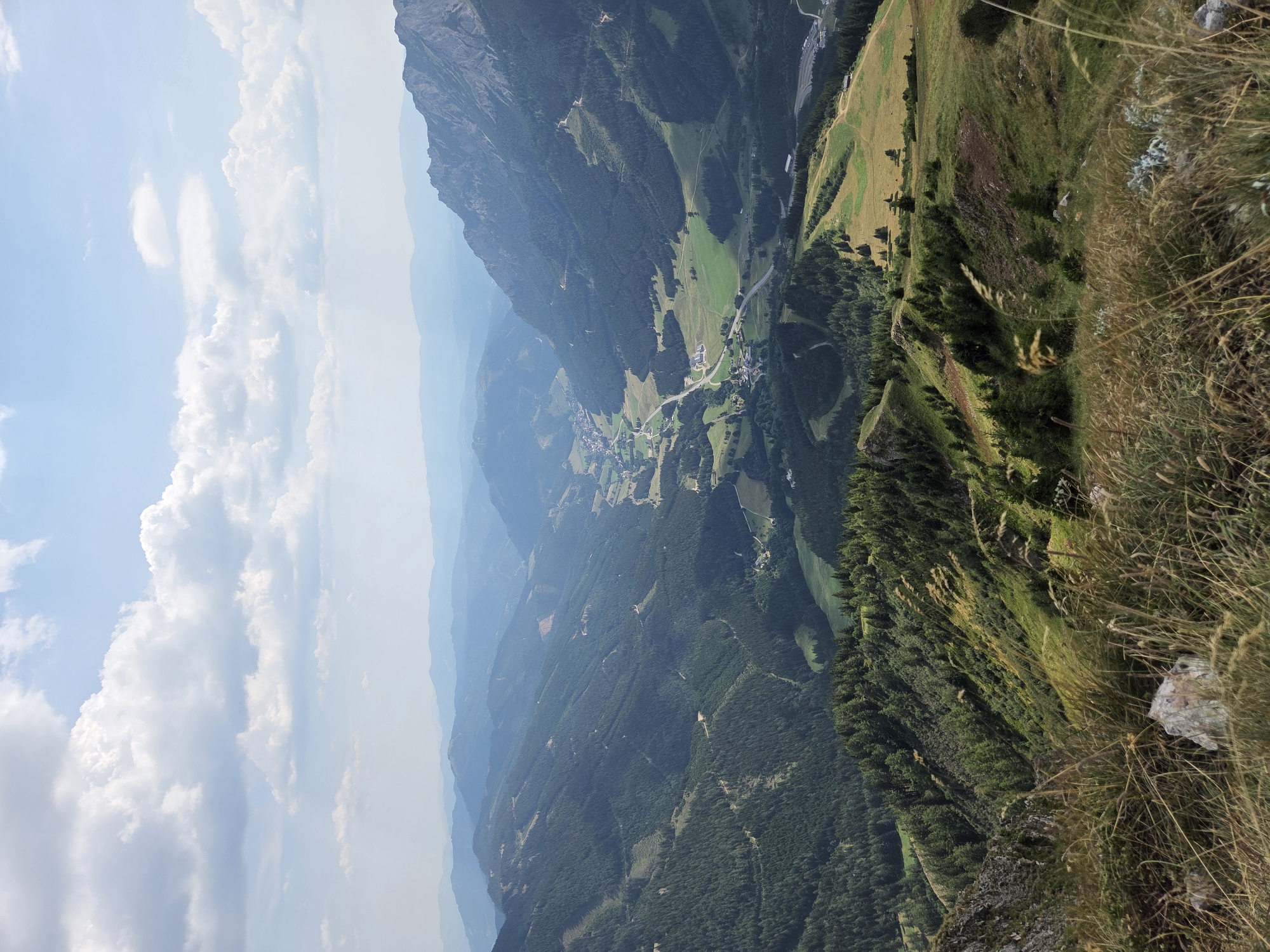 The image captures a sweeping panoramic view of a mountainous valley, likely taken from an elevated vantage point in or near Vordernberg, Austria. The scene is dominated by lush green, tree-covered slopes that recede into the distance, forming layers of mountains against a pale blue sky dotted with fluffy white clouds. A small village or town is nestled in the valley floor, characterized by scattered buildings and a visible road network winding through the landscape. The foreground of the image features dry grasses and rocks, suggesting a natural, undeveloped area. The overall impression is one of a serene and expansive natural landscape with a human settlement integrated into it.