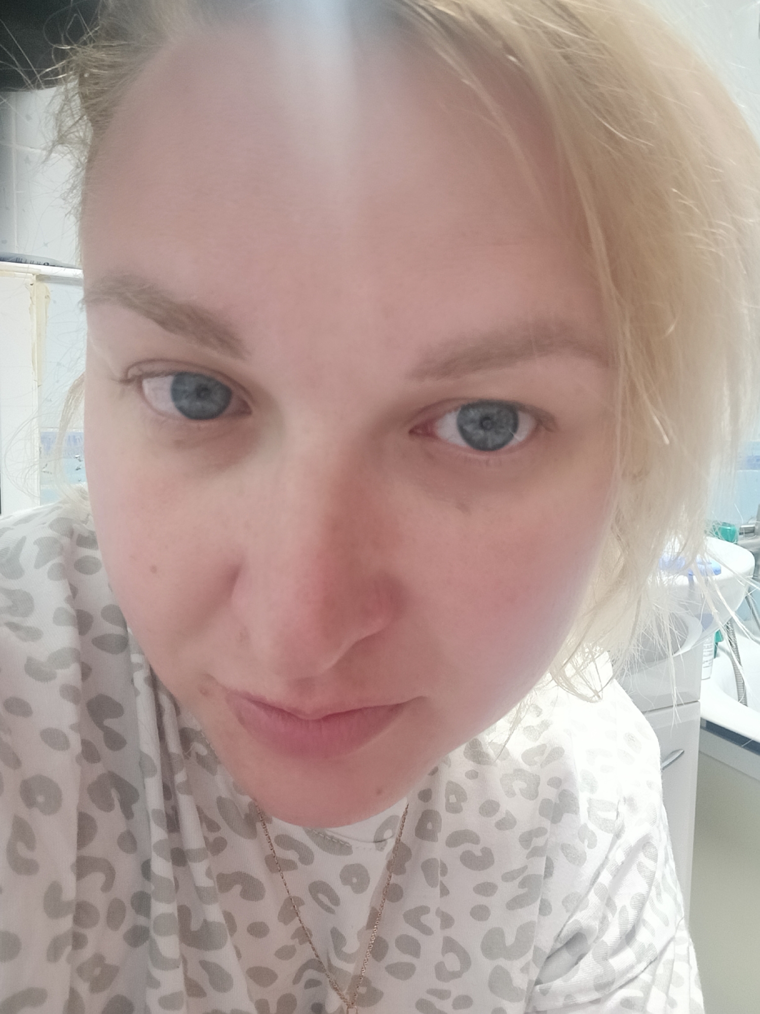A close-up, selfie-style photo of a fair-skinned woman with blonde hair and blue eyes. She's wearing a light-colored top with a grey leopard print. A thin gold chain necklace is visible around her neck. The background appears to be a medical or clinical setting, evident from the sink, countertop, and medical equipment slightly visible behind her. The location is unknown.