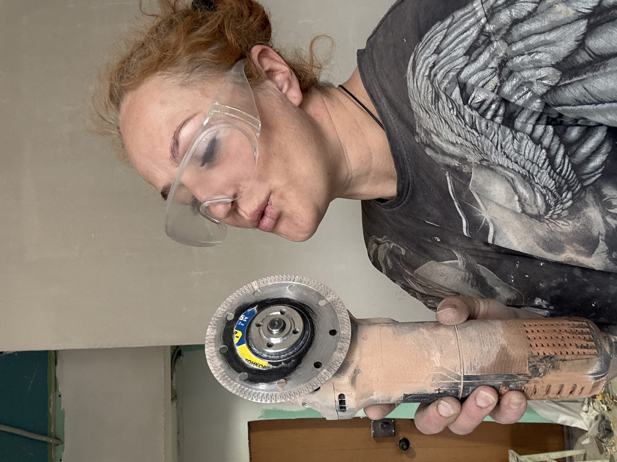 A woman wearing safety glasses is holding a power tool with a circular blade. The tool appears to be used, covered in a light brown dust. The woman is wearing a dark grey t-shirt with a large, light grey graphic. The background includes a wall painted light grey with sections of teal and possibly yellow. A wooden door is also visible. There is no indication that the image was taken in Chaikovsky, Russia.