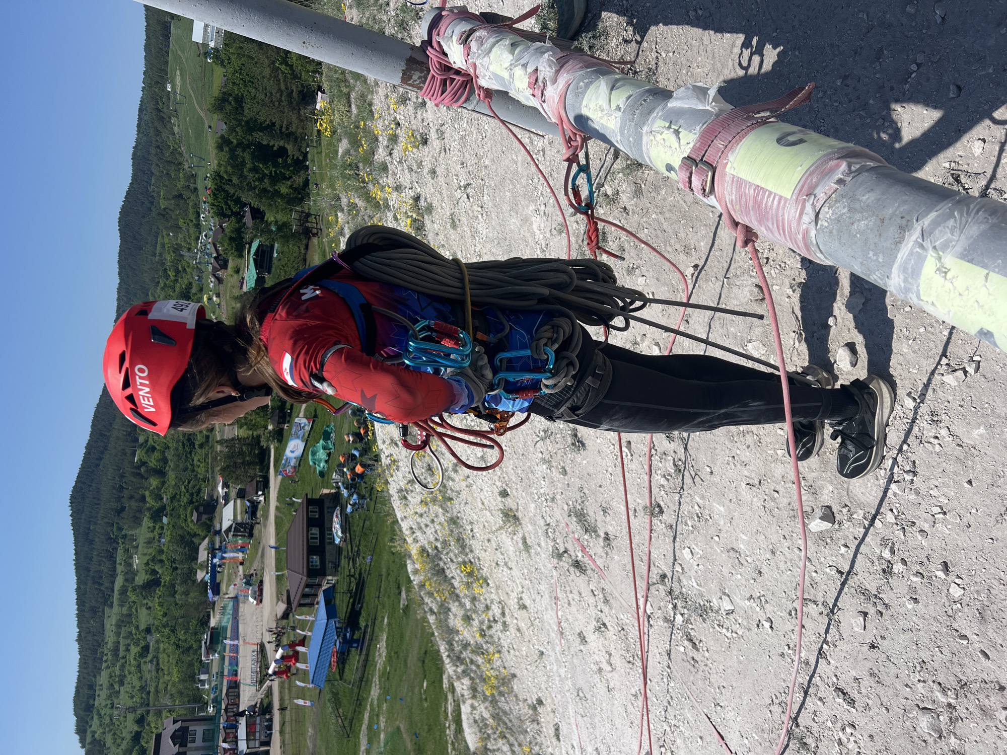 A person wearing a red helmet, a red and blue jacket, black pants, and black shoes is rappelling down a gray pipe. The person is connected to the pipe with ropes and carabiners. A town with buildings, trees, and a dirt road is in the background. The pipe is wrapped with pink straps and yellow tape. The ground is light gray and covered with small rocks and yellow flowers. The photo was taken outdoors during the day. The location is Khvalynsk, Russia.