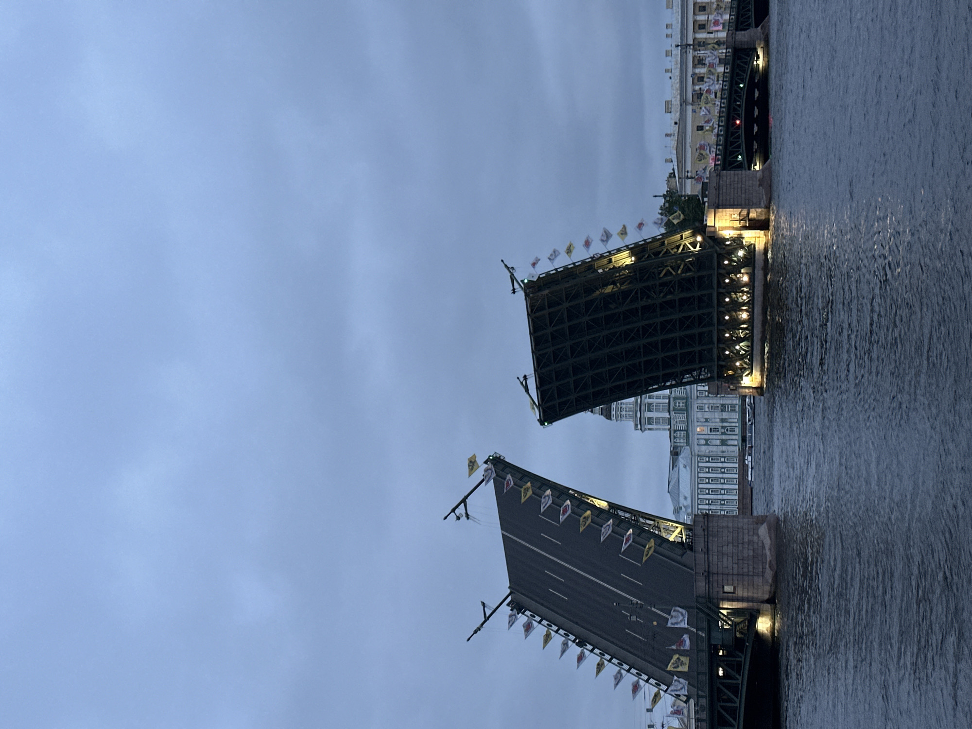 A drawbridge is raised in the city of St. Petersburg, Russia, over the Neva River at dusk. The sky is overcast, and the water is dark and choppy. The drawbridge consists of two large sections that are angled upwards, revealing their internal structural framework and undersides. The roadway on the left section is visible, with white lines marking lanes. Both sides of the bridge are adorned with numerous small flags of various colors, including yellow, red, and white, which flutter in the wind. Numerous lights illuminate the underside of the bridge sections. On the riverbank behind the bridge, several neoclassical buildings are visible. The most prominent is a large, pale building with a dome and many windows. Other buildings, some adorned with more flags, line the embankment. The scene depicts a moment of transition, with the bridge raised to allow passage for vessels on the river.