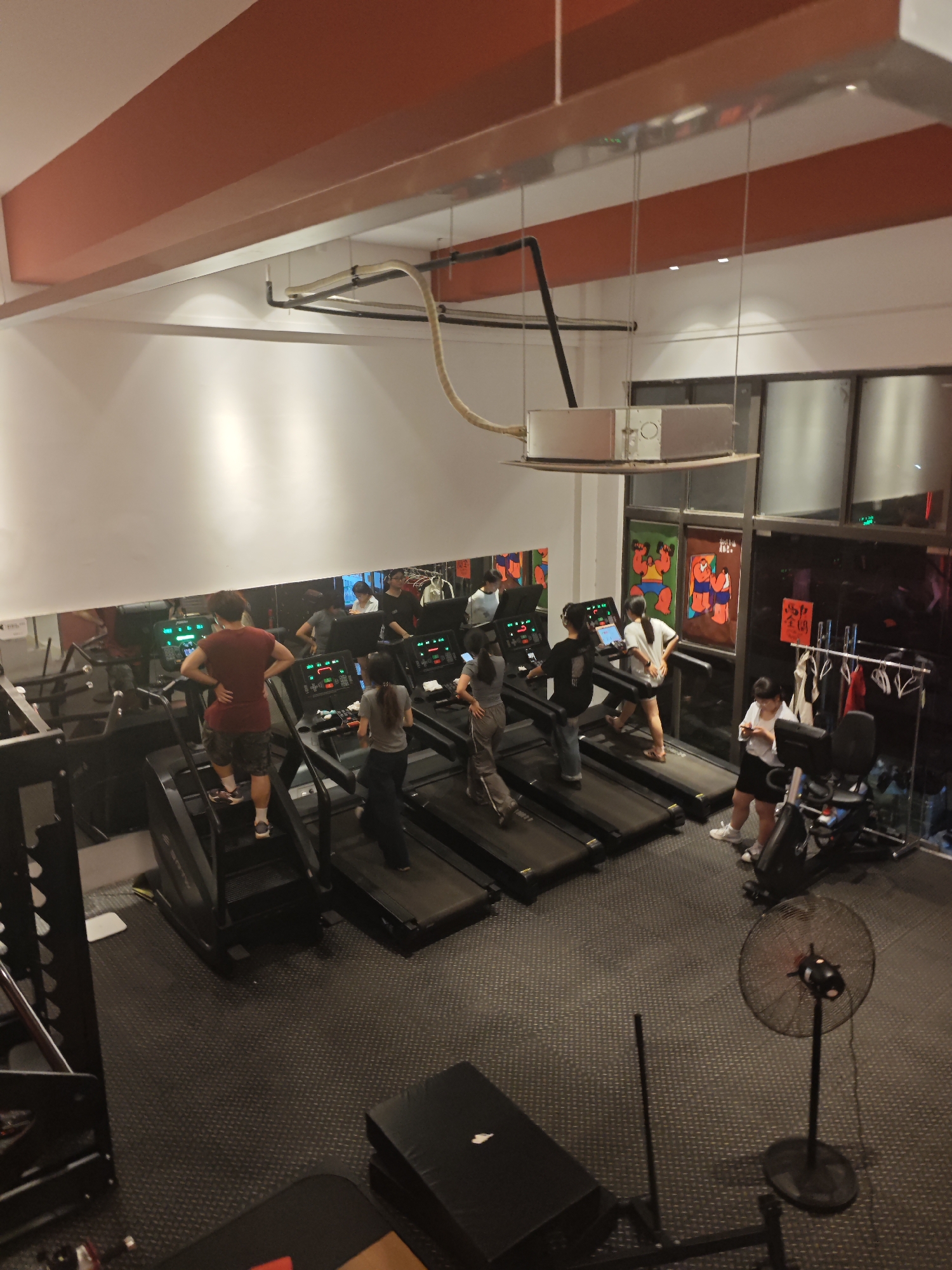 The image provides an elevated view of an indoor gym or fitness center, likely situated in Chongqing High-tech Zone, China. The scene depicts several individuals, primarily young adults, engaged in various workout activities.
In the foreground and mid-ground, a row of treadmills and a stair-climbing machine are in use. A man in a red sleeveless shirt stands on the stair-climber, facing away from the camera. Alongside him, several people are on treadmills, either walking or running. One woman is seen standing near an exercise bike on the right, occupied with her mobile phone. Reflections of people and equipment are prominently visible in the large mirrors lining the left wall, which creates an impression of a larger space.
The gym's interior features dark, textured rubber flooring. The walls are predominantly white, contrasted by a distinct orange-red beam that spans across the upper ceiling/wall area. Overhead, an industrial-style ventilation system with exposed pipes and a rectangular metal unit is suspended. Artificial lighting illuminates the area. On the right side of the gym floor, a large black standing fan is visible. Behind the exercise bike, large glass panels reveal a section of a wall adorned with colorful, stylized artwork depicting figures, possibly reflecting local artistic or cultural influences. Some red text, potentially Chinese characters, is faintly discernible near the windows but is illegible from this distance. Clothes are hung on racks near these windows. The atmosphere suggests a typical active gym environment.