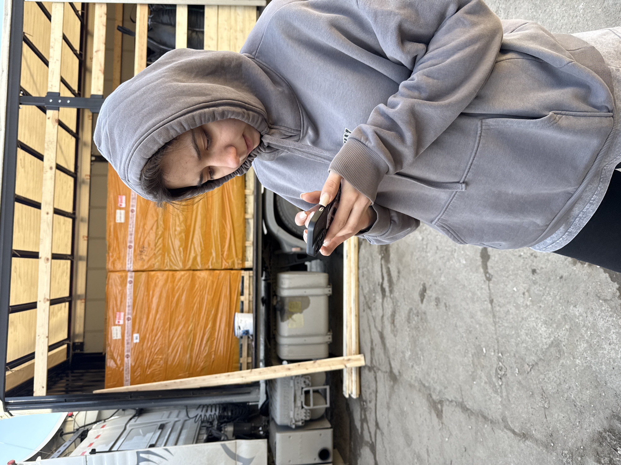 A person wearing a gray hooded sweatshirt is standing next to an open trailer truck. The trailer contains boxes wrapped in orange plastic. The person is looking down at a smartphone held in their hands. The environment appears to be an outdoor loading area, with a concrete ground visible.