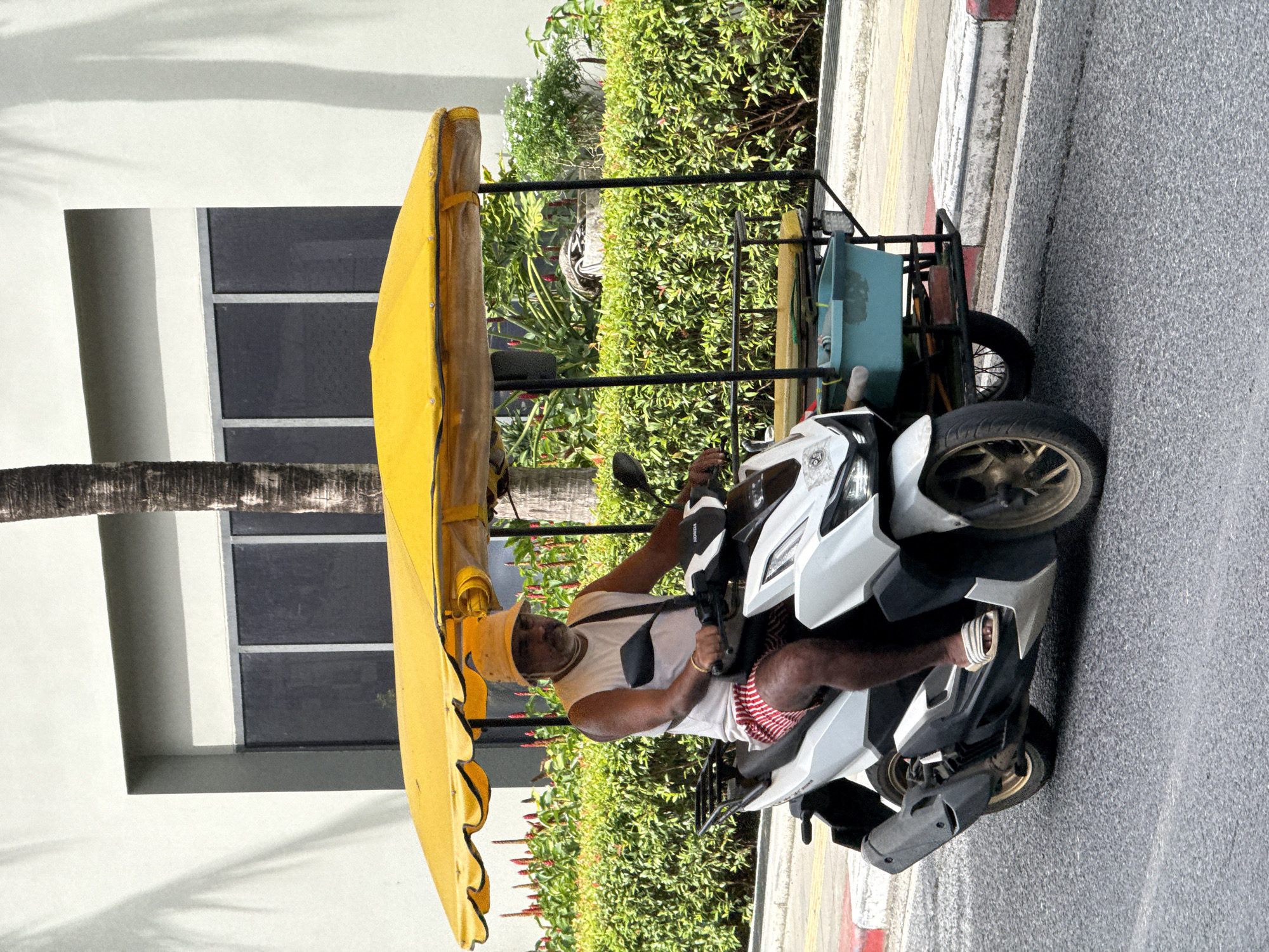 A middle-aged man wearing a white sleeveless shirt, red and white striped shorts, a yellow bucket hat, and white striped sandals rides a white three-wheeled scooter equipped with a yellow awning. The scooter has a turquoise bin and a metal rack attached to the rear. The man is on a paved road with red and white curbs. In the background, there is a building with gray siding and dark windows, a palm tree, and lush green bushes with red and white flowers. The scene is set in Karon, Thailand, under natural daylight.