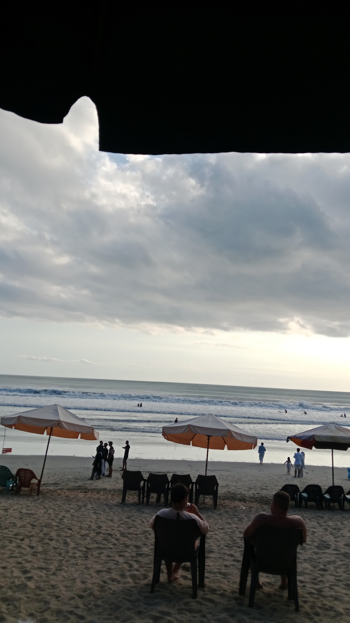 The image shows a beach scene in Kuta, Indonesia. The sky is overcast with dramatic clouds. In the foreground, there is a sandy beach with several empty chairs and umbrellas. Two men are seated in chairs, facing the ocean. Further down the beach, closer to the water, a group of people are standing and walking. The ocean has moderate waves, and several surfers are visible in the water. The overall atmosphere appears to be a late afternoon or early evening on a cloudy day at the beach.