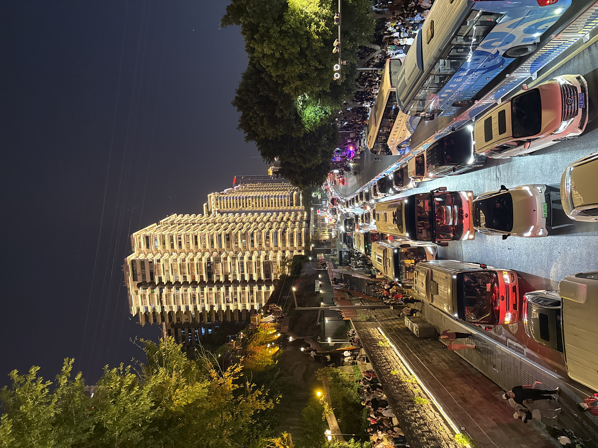This night-time aerial perspective captures a bustling urban scene in Yuzhong District, China, showcasing a vibrant mix of heavy traffic and large crowds. The image is dominated by a multi-lane road on the right, filled with a dense line of vehicles, including buses, cars, and vans, indicating significant traffic congestion. Several vehicles display license plates starting with "渝A", characteristic of Chongqing, and a blue and white bus prominently features "重庆公交" (Chongqing Public Transport) in Chinese characters.
Alongside the road, a wide sidewalk is packed with numerous people, forming a lively crowd. Further to the left, a tall, modern building with many illuminated windows or balconies stands brightly lit against the dark sky, suggesting residential or commercial use. Below this building, a lower pedestrian promenade or pathway is also populated with people, some walking, others standing or seated. Trees and green vegetation are scattered throughout the scene, bordering the road and the lower path.
The sky is dark, affirming the night-time setting, with power lines faintly visible. The numerous streetlights and vehicle headlights, along with the illuminated buildings, cast a warm glow across the scene. The overall impression is one of a busy, energetic urban environment, possibly a popular gathering spot or a route experiencing peak activity. The clear weather allows for good visibility of the detailed scene.