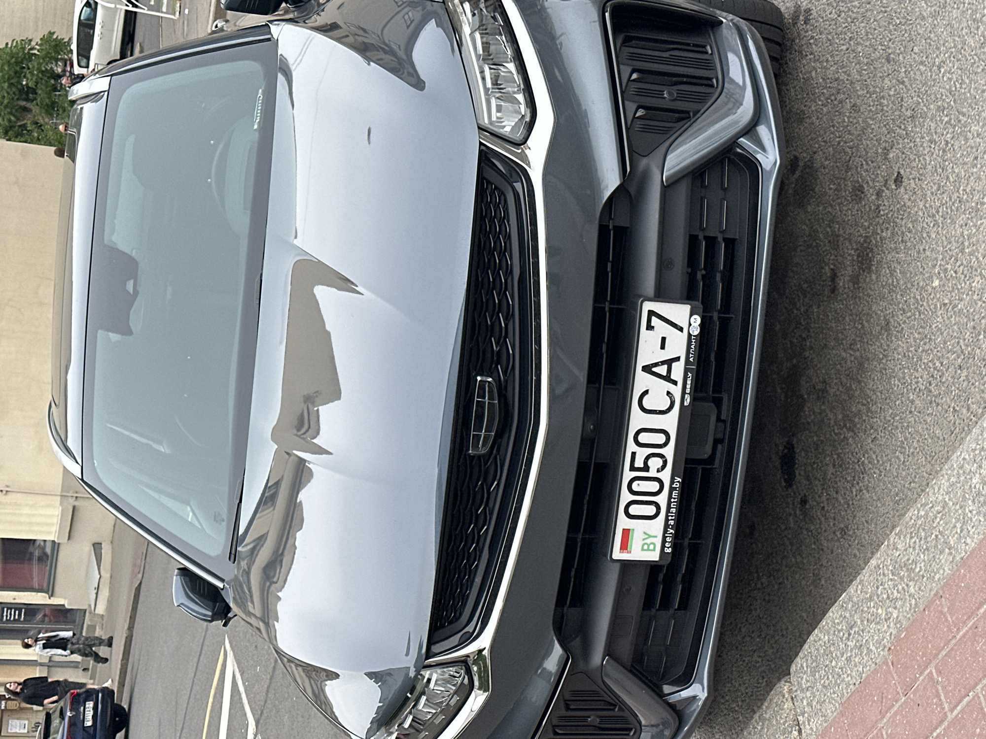 A dark grey Geely SUV is centrally parked on an asphalt street, adjacent to a concrete curb with a red brick edge. The vehicle displays a Belarusian license plate: "BY 0050 CA-7". The license plate frame includes "geely-atlantm.by" and "АТЛАНТ-М" text. The car's front grille features the Geely logo, flanked by LED headlights and a stylized lower bumper with vertical vents. Reflections of surrounding buildings and the sky are visible on the car's hood and windshield.
In the background, two individuals are walking on a light-colored sidewalk. The person on the left has long dark hair, wearing a black t-shirt and dark pants. The person on the right has shorter dark hair, wearing a light jacket over a dark top and camouflage-patterned pants. Behind them stands a multi-story building with light-colored walls and windows, indicating an urban setting. Further down the street, another dark vehicle (partially visible on the left, with a Belarusian license plate starting "711AB-") and a white minivan (on the right) are parked. Green foliage from trees is visible above the buildings on the right.
The scene depicts an urban street in the city of Minsk, Belarus, as evidenced by the Belarusian license plates and the "Atlant-M" dealership reference, which is a prominent automotive group in the country.