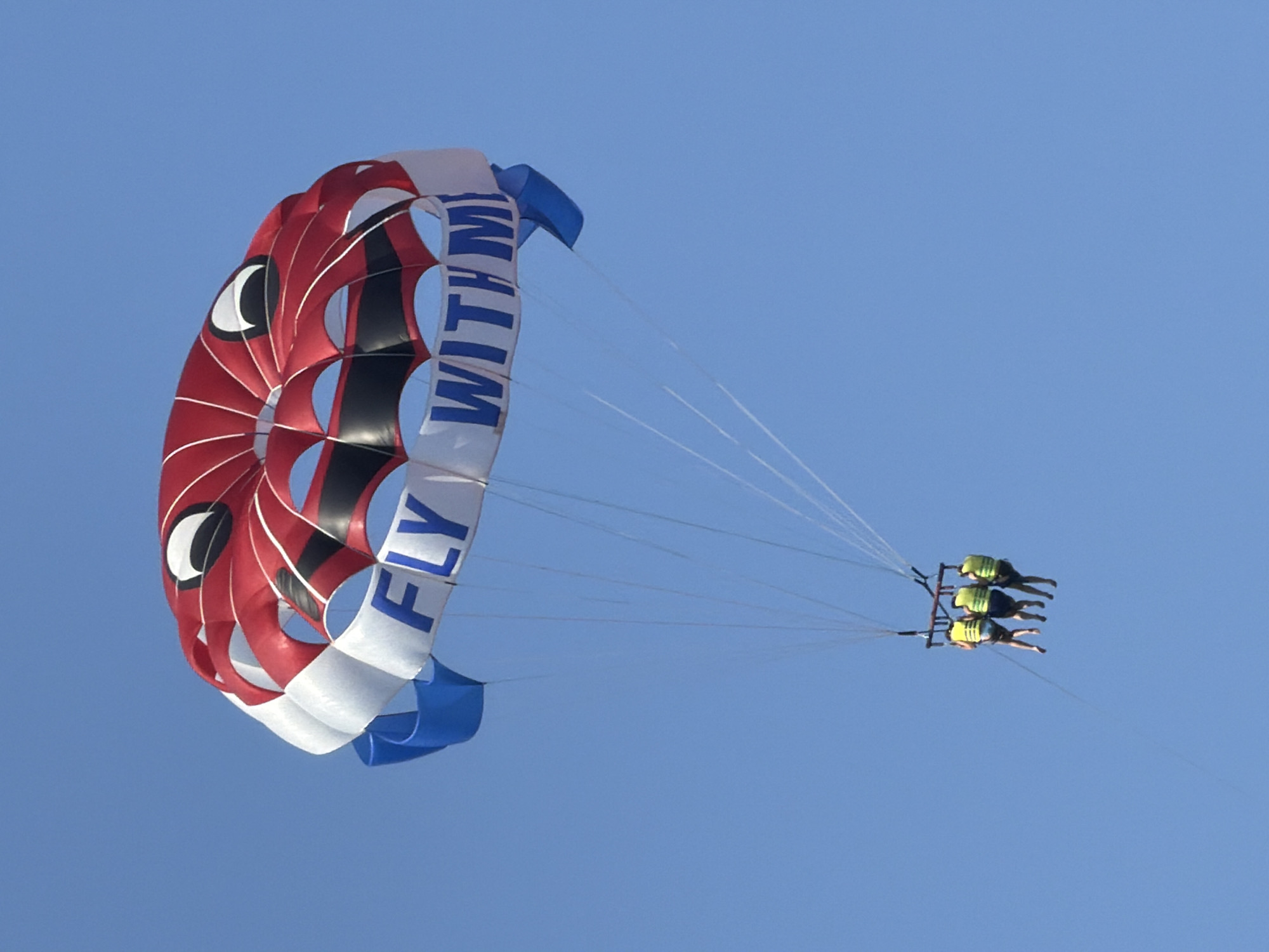 This media file shows three people parasailing against a clear blue sky. The parasail is predominantly red with white and black accents, and a blue section at the bottom. Large white letters spelling "FLY WITH ME" are visible on the side of the parasail. The three people are suspended beneath the parasail, each wearing a yellow life vest. They appear to be enjoying the activity, with their bodies positioned to face forward as they are pulled through the air. The lighting suggests it is daytime, likely a sunny day given the bright blue sky and clear visibility. The image is taken from a low angle, looking up at the parasail and the people. The focus is on the parasailing activity, with the vast expanse of the sky serving as the backdrop.