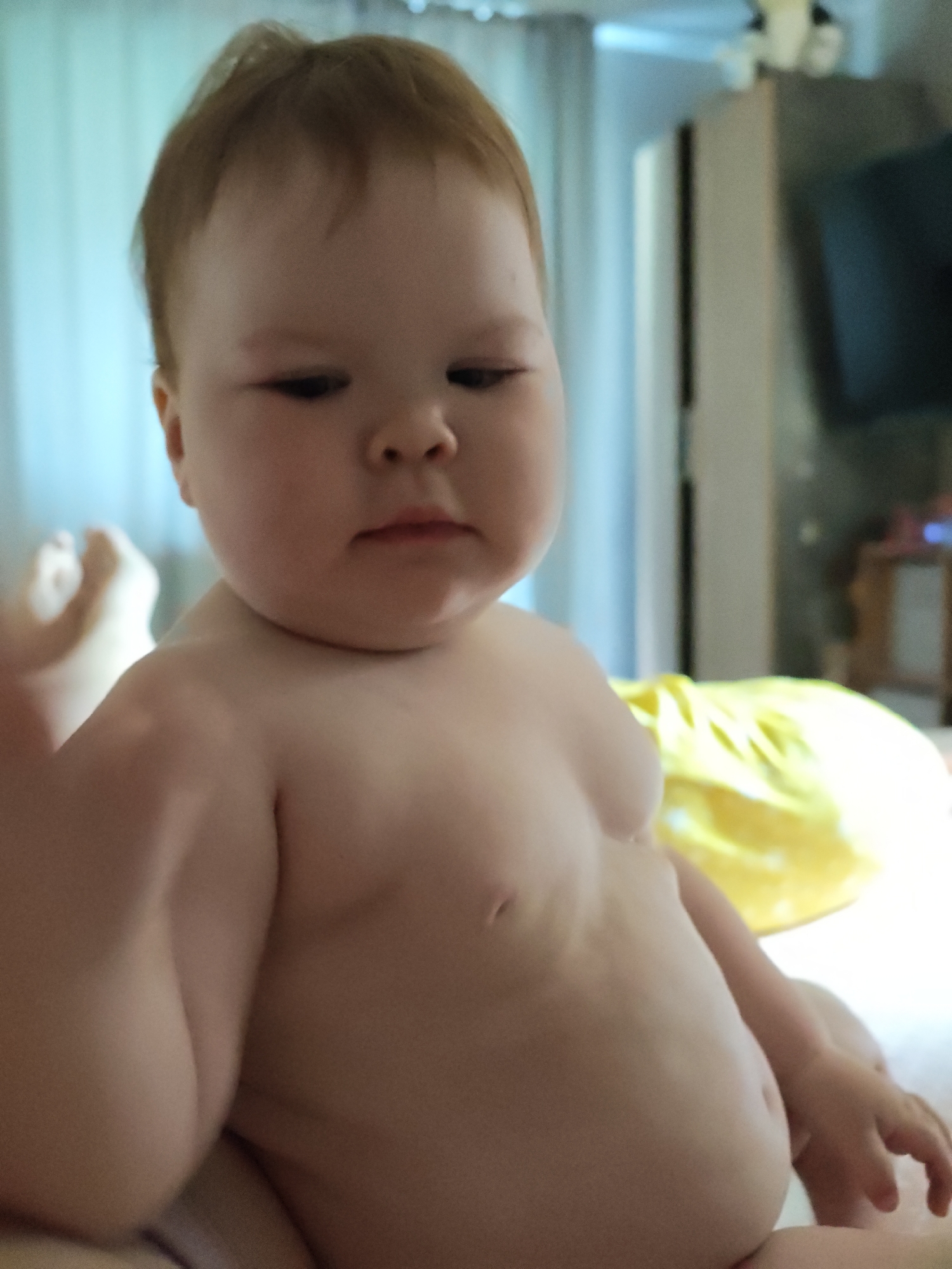 A baby with reddish-brown hair is positioned in the center of the frame, looking down and slightly to the left. The baby is shirtless.
The scene appears to be indoors, potentially a bedroom or living space, with blurred curtains and a TV. A partially visible leg and foot and a yellow textile are also present in the frame.
There is no information to identify that the picture was taken in the city of Chaikovsky, Russia.