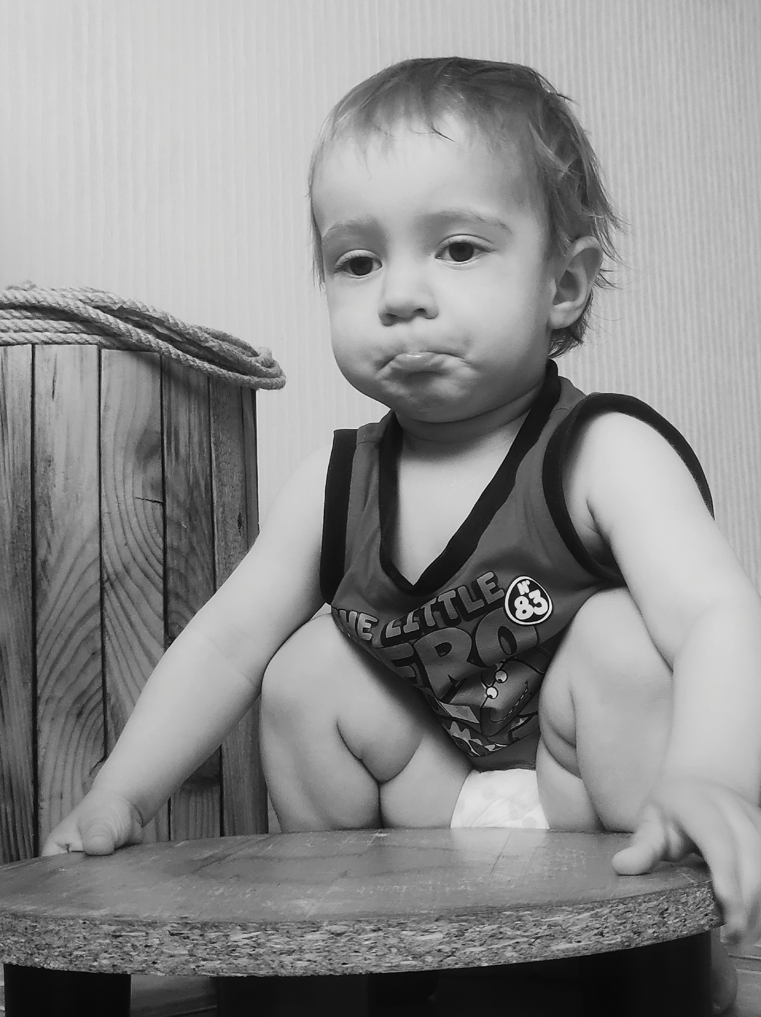 The image is a monochrome photograph of a young child, possibly a toddler, sitting on a round, possibly wooden, surface supported by black legs. The child is wearing a tank top with a graphic print on the front. The child's expression appears to be pouting or displeased. To the left of the child is a wooden barrel or container with a rope coiled on top. The background is a plain wall with a subtle vertical pattern. The image was captured in Chaikovsky, Russia.