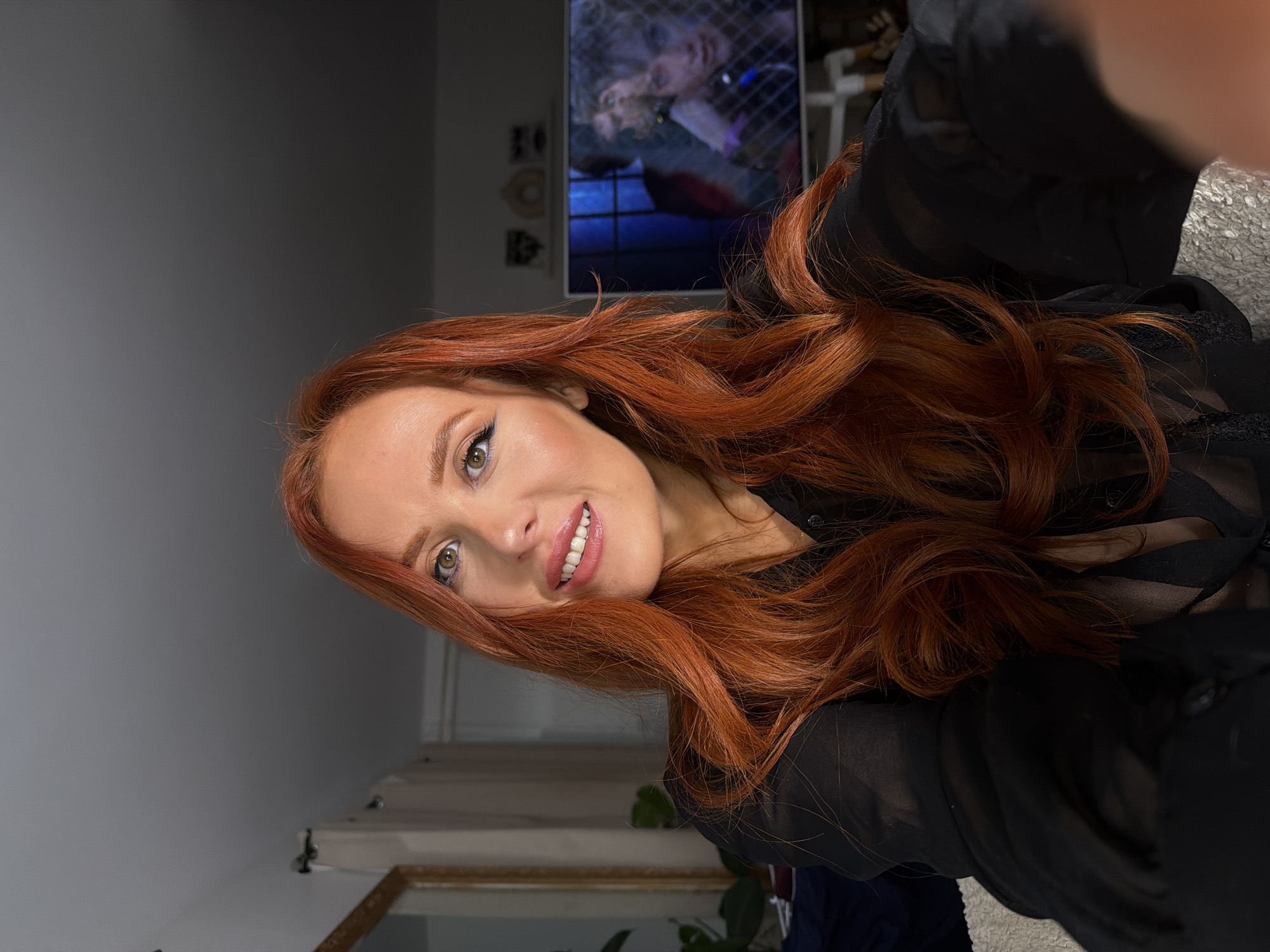 A selfie-style photograph shows a woman with red hair, fair skin, and light eyes smiling. She is wearing a black sheer top. In the background is a television screen showing a blurred image of a person. The background also includes a wall that is a light grey color. The image appears to be taken indoors. There is no indication that the location is in Paris, France.