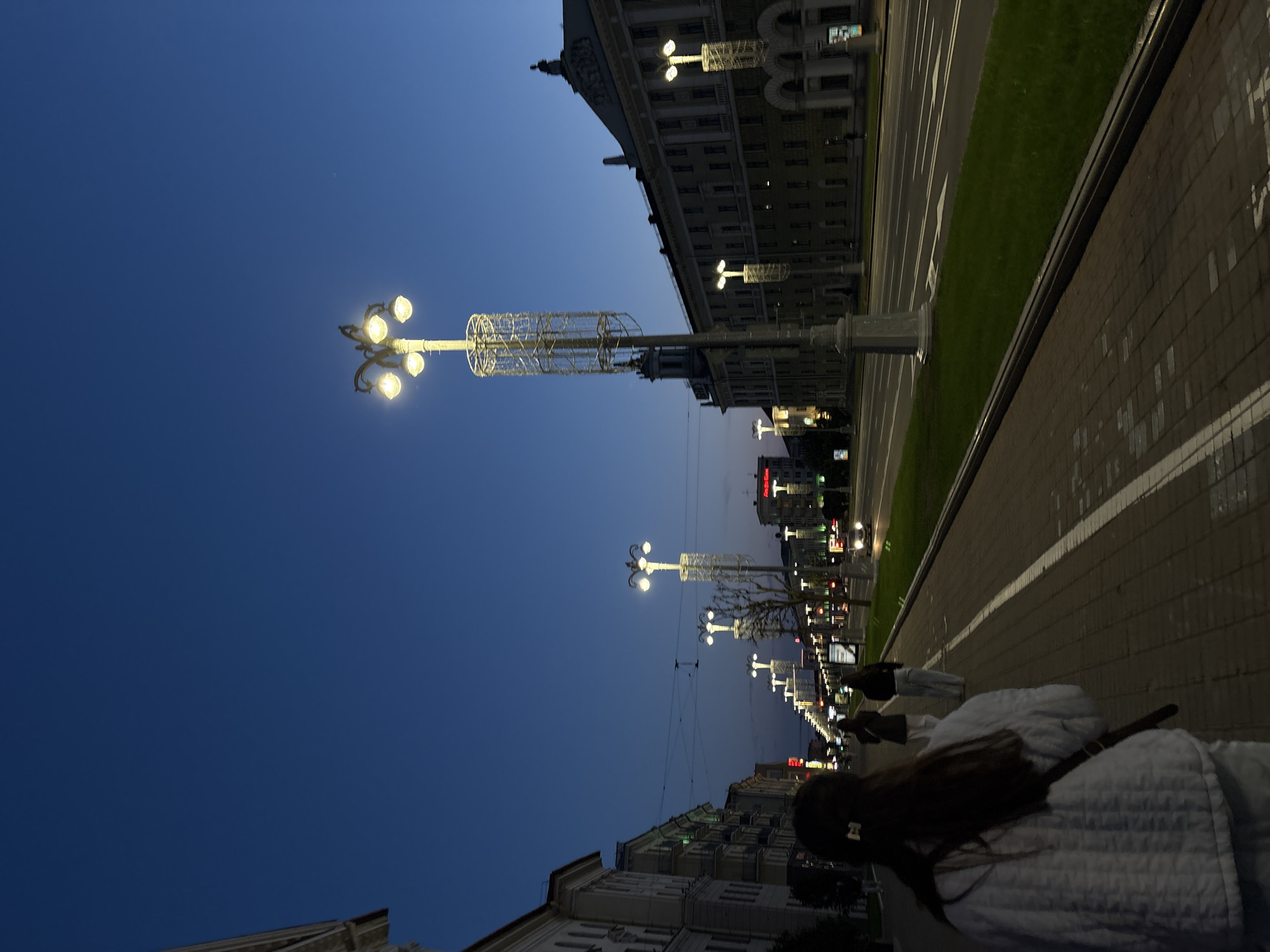 The image depicts a nighttime street scene in Minsk, Belarus. A row of ornate streetlights illuminates the boulevard, running parallel to the buildings and a broad sidewalk. The streetlights have a distinctive design with multiple globe-shaped lights. A person wearing a light-colored quilted jacket with long brown hair and a black backpack is visible in the foreground. Other individuals are walking further down the sidewalk. Tall buildings line the street with visible architectural details. The sky is a deep twilight blue. The scene is oriented vertically.