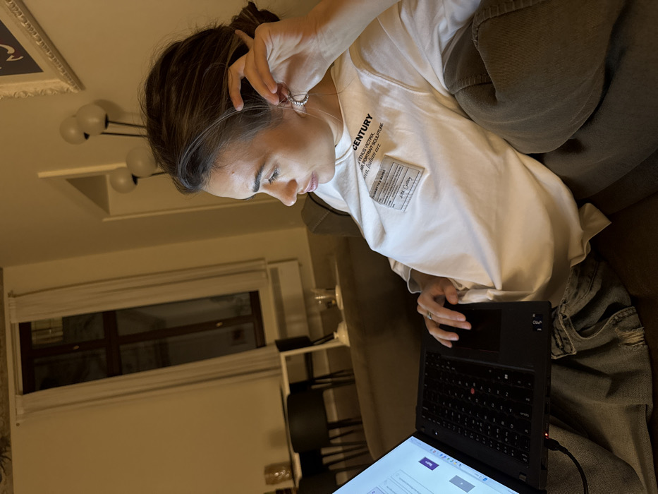 A single female is depicted seated on a sofa indoors during nighttime, engaged with a laptop. The woman, appearing in her late teens or twenties, has brown hair styled back and wears a white t-shirt featuring prominent text, including "CENTURY" and "VENUS VICTRIX," and light-colored pants. A silver-colored ring is visible on her left ring finger. She is looking intently at the laptop screen, which displays a light-colored interface with text and a "Next" button. Her left hand is positioned near the laptop's trackpad, while her right hand is raised, touching a silver hoop earring in her right ear. The black laptop, likely a Lenovo ThinkPad, is connected via a red-tipped charging cable.
The interior space is illuminated by warm, ambient lighting. In the background to the left, a dining area is visible with a dark table and chairs. Beyond this, a tall window fitted with white curtains reveals a dark exterior, consistent with nighttime. A decorative object, possibly a vase, stands on a surface near the window. To the right, a floor lamp with three spherical white shades and a partially visible framed picture are present. The scene is located in the city of Venice, Italy.