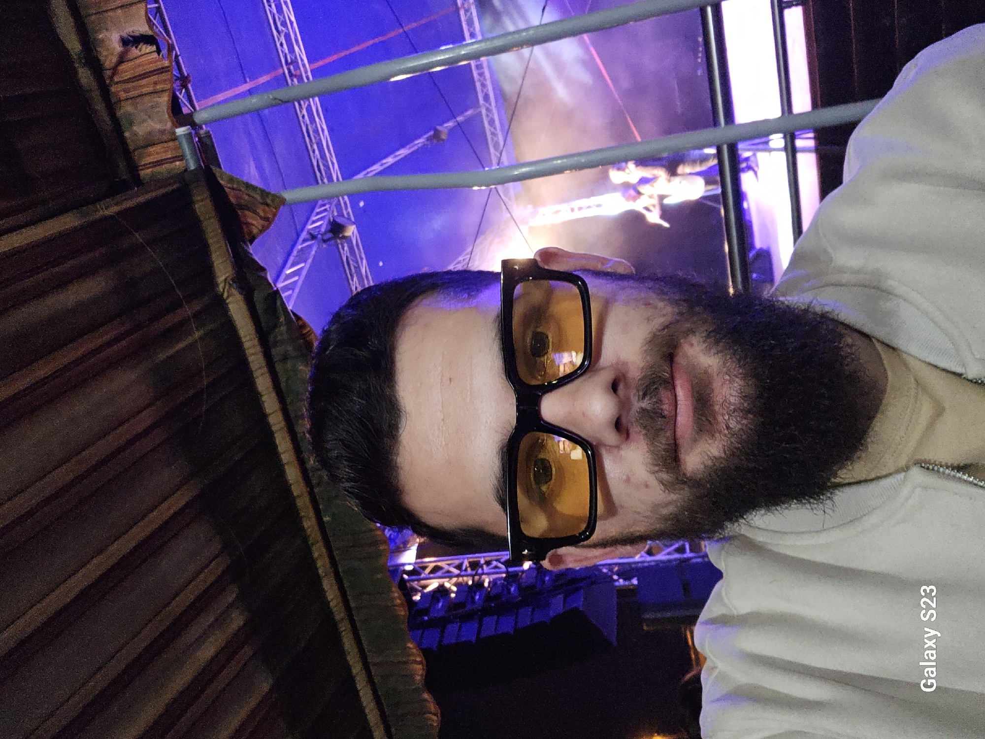 The image is a selfie of a man with a beard and glasses, likely taken at a concert or event. The background shows a stage with lighting and what appears to be performers. Structural elements such as beams and supports are visible, suggesting an indoor or covered venue. The photo was taken with a Samsung S23 phone. The city of Sliven, Bulgaria, is not apparent from the image and cannot be confirmed without additional information.