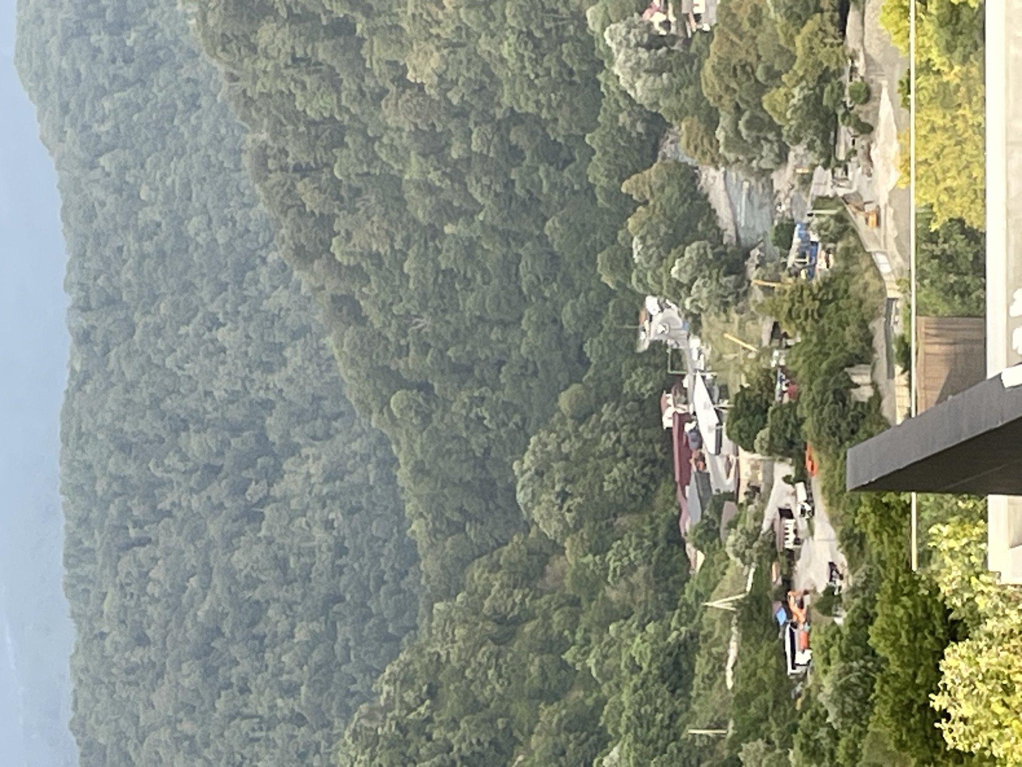 The image captures an aerial view of a construction site nestled within a lush, forested mountain valley. In the foreground, the edge of a balcony railing and some greenery are visible. The midground showcases a scene of activity: several construction vehicles, including excavators and trucks, are positioned on a dusty road and amidst piles of building materials. Buildings, some with red roofs, are scattered throughout the area. A river winds its way through the valley, bordered by dense trees. The background is dominated by a vast expanse of verdant, undulating hills covered in thick forest. The overall impression is a landscape undergoing development within a natural, mountainous setting, likely in the vicinity of Baranovka, Russia, given its characteristic geography.