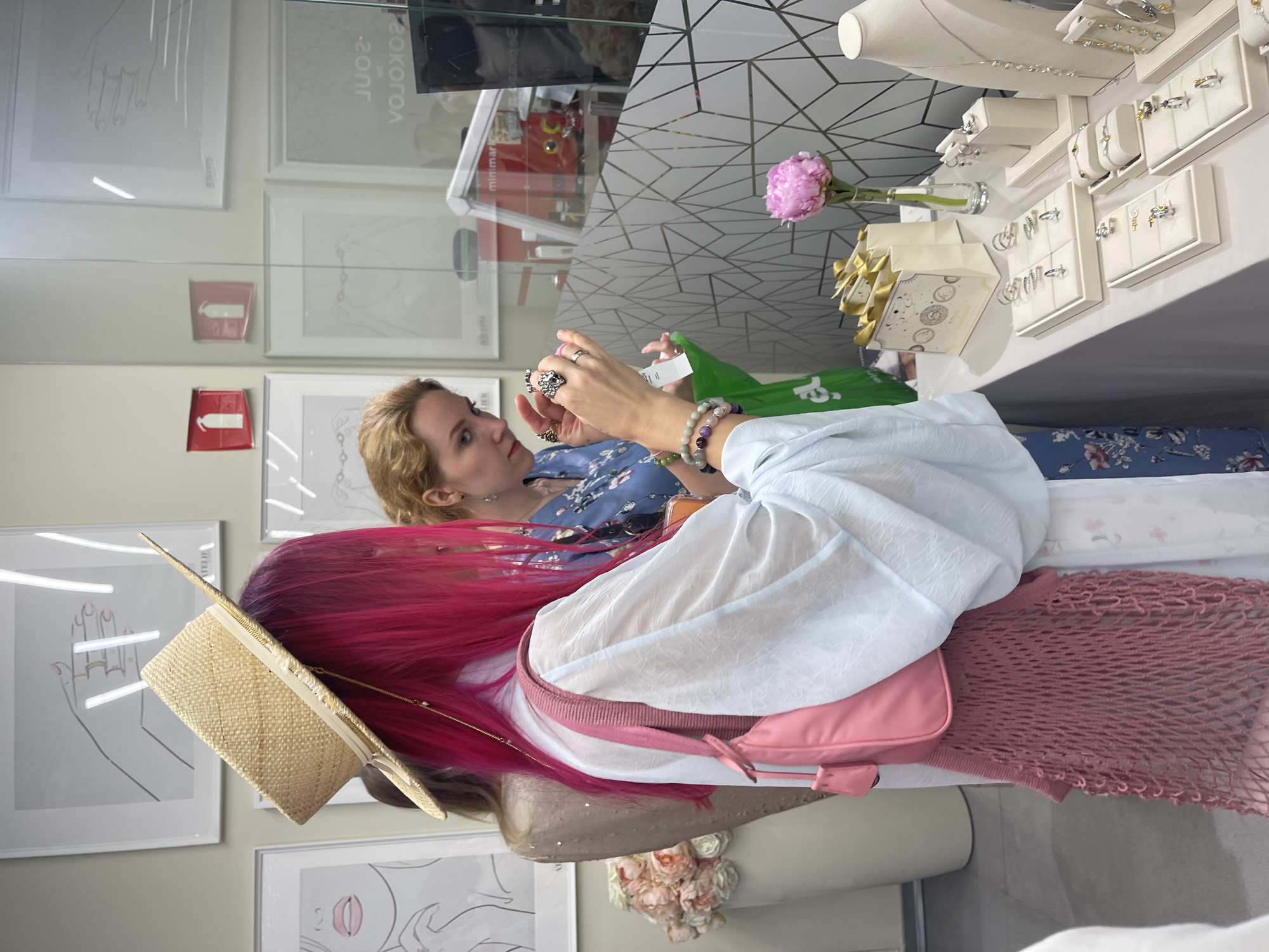 The image depicts an indoor scene at a jewelry retail location, likely in Moscow, Russia, indicated by the reversed reflection of "SOKOLOV," a prominent Russian jewelry brand, on a glass partition.
In the foreground, a person with vibrant fuchsia-pink hair, wearing a straw boater hat with a white ribbon, a light-colored translucent blouse, a pink shoulder bag, and a pink mesh layer underneath, stands with their back mostly to the viewer, looking down at a jewelry display. This individual's left arm is visible, adorned with multiple beaded bracelets in shades of purple, pink, white, and green.
To the right, a woman with light blonde hair, wearing a blue floral patterned dress, intently examines a silver ring on her right hand. Her left hand holds a small white tag. She has additional rings on other fingers.
The central feature is a white display table showcasing various silver and possibly gold-accented jewelry, including rings, necklaces, and bracelets, presented on white display trays and stands. Behind these displays, several small white gift bags with gold handles and celestial designs are arranged. A single pink peony in a clear glass vase sits on the table.
The background features light-colored walls adorned with framed, minimalist line art drawings of hands, faces, and abstract forms. Two recessed red wall compartments, possibly indicating fire safety equipment covers, are also visible. A tall white planter containing light pink and white artificial or dried flowers is on the far left. A glass partition, partially covered with a grey geometric pattern, separates the display area from a background space where "minimart" is faintly reflected along with "SOKOLOV" and "SOUL" in reverse. The overall environment suggests a well-lit, contemporary retail setting.