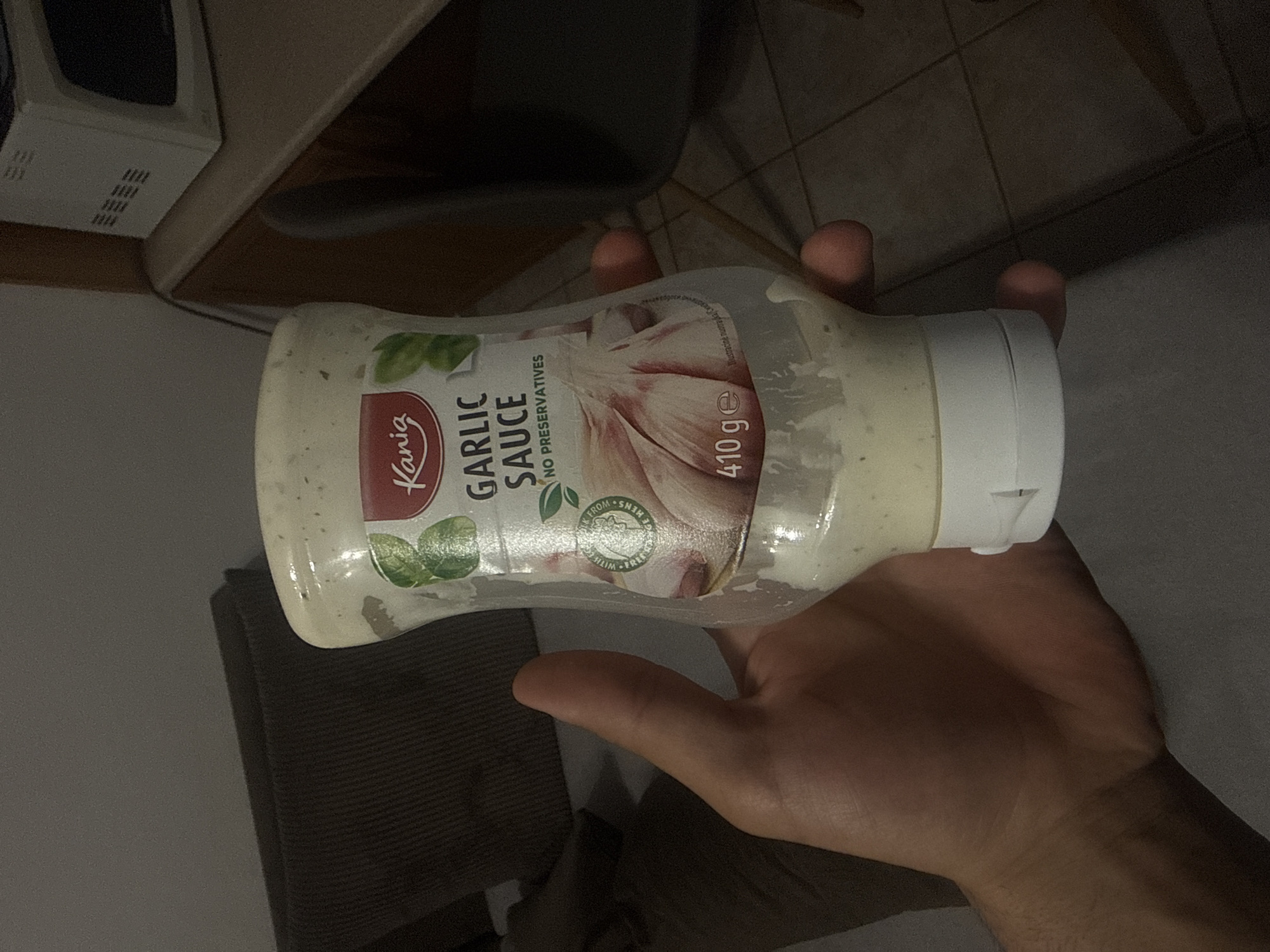 The scene depicts an indoor residential environment in Varna, Bulgaria. A human right hand, exhibiting a light to medium skin tone, is holding a partially depleted, clear plastic squeeze bottle of Kania Garlic Sauce. The bottle's label, oriented towards the viewer, displays "Kania" branding, "GARLIC SAUCE," "NO PRESERVATIVES," an illustration of garlic bulbs, and a net content of "410 g e." Residual white, creamy sauce containing green specks adheres to the interior walls of the bottle, which features a white flip-top cap. To the left of the hand, a dark brown/grey ribbed fabric cushion or upholstery is visible, positioned against a light grey wall. To the right, a light brown wooden countertop supports a white microwave oven with a dark door and side vents. A dark, possibly metal, chair component is partially discernible beneath the countertop, and the floor is tiled with light brown square tiles. A dark cable or wire runs vertically in the background on the right. The ambient lighting is dim, suggesting a low-light condition.