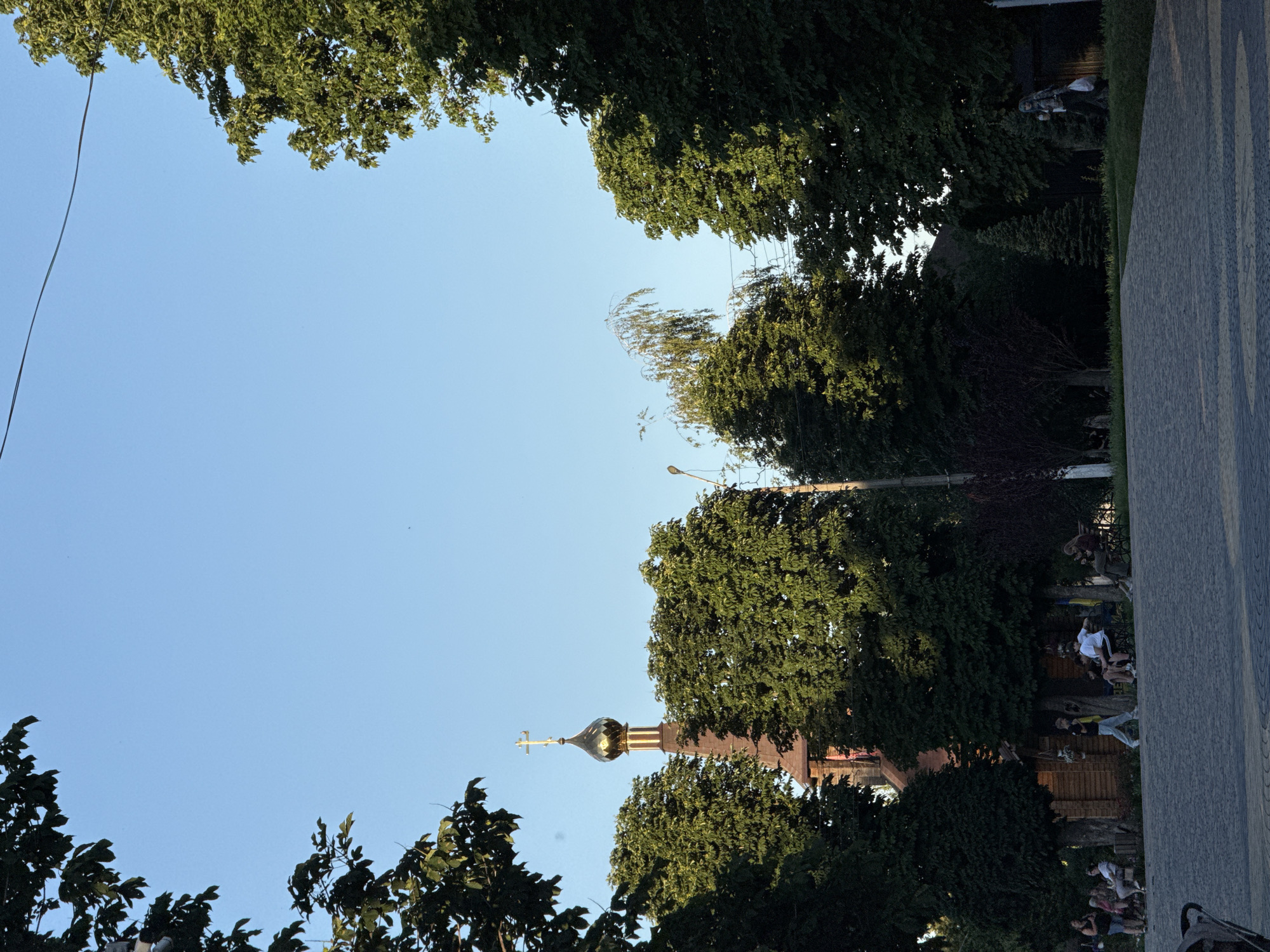 In a city park under a clear blue sky, a wooden church with a golden dome and a cross stands amidst lush green trees. A paved walkway leads to the church, where several people are seated on benches, some conversing, others looking at their phones. A few individuals are walking along the path, and one person is strolling past a lamppost on the right side of the frame. The scene is bathed in warm sunlight, casting gentle shadows across the park.