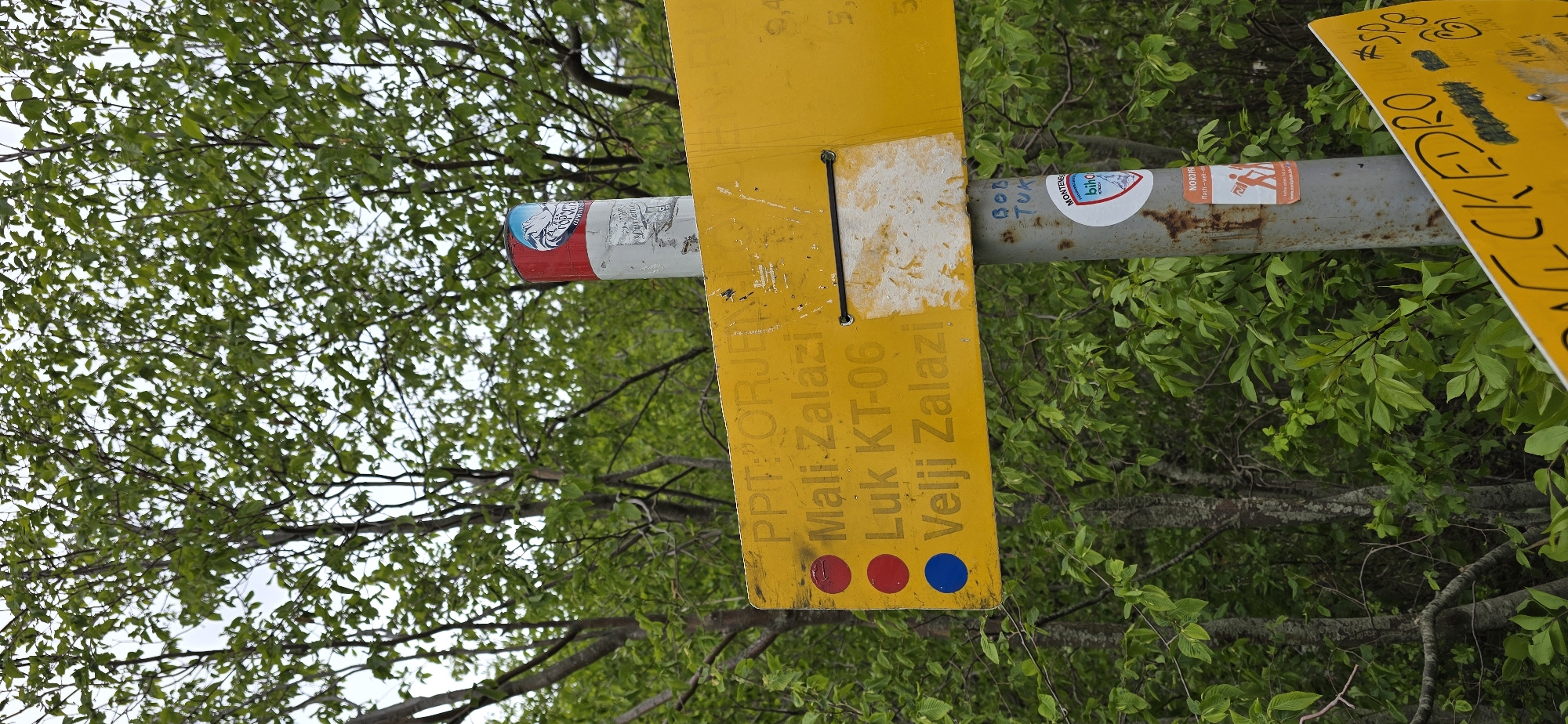 Here is a description of the image:
The image shows a trail marker, likely in a wooded area within the vicinity of Kopito, Montenegro. The trail marker, bright yellow in color, is attached to a metal pole. The marker displays information about trails: "PPT: ORJEN Mali Zalazi Luk KT-06 Velji Zalazi". Three colored dots, two red and one blue, are visible at the bottom of the sign. Stickers and signs are affixed to the metal pole. The background is predominantly composed of green foliage, suggesting a wooded environment.