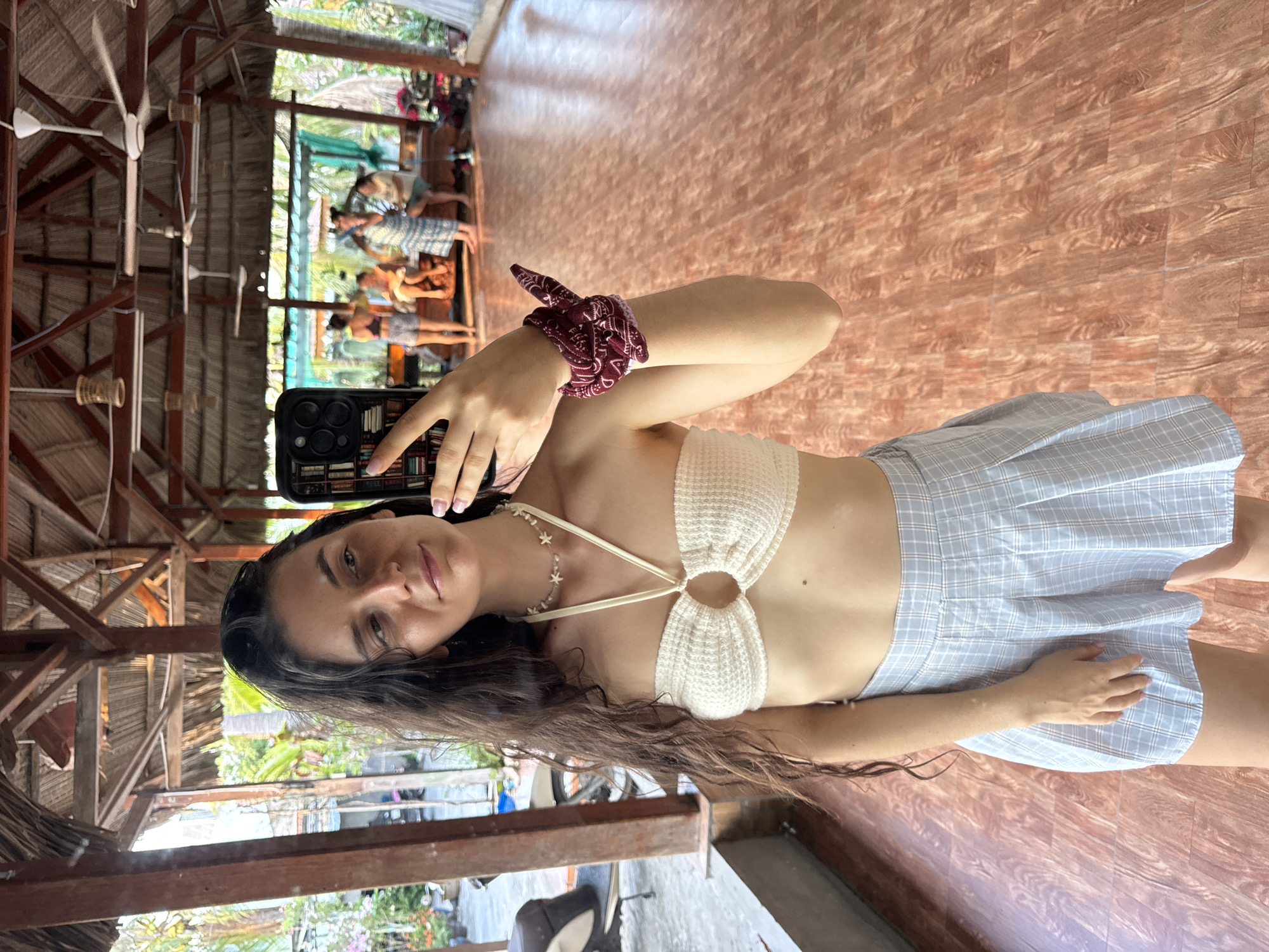 A woman is taking a selfie in what appears to be an outdoor restaurant or open-air structure. The woman has long dark hair and is wearing a light-colored crocheted top and a blue and white plaid skirt. She has a patterned red and white hair tie or bandana around her wrist. She is holding a smartphone with a decorative case to take the photo.
The location has a rustic, tropical feel. The structure is constructed of wooden beams and has a thatched roof. In the background, there are other people seated at tables, indicating a dining environment. The flooring is made of wood planks. The structure seems to be open to the outside, with glimpses of tropical plants and vegetation visible. Based on the tropical environment and the architectural style, it is likely that the photo was taken in Mũi Né, Vietnam.