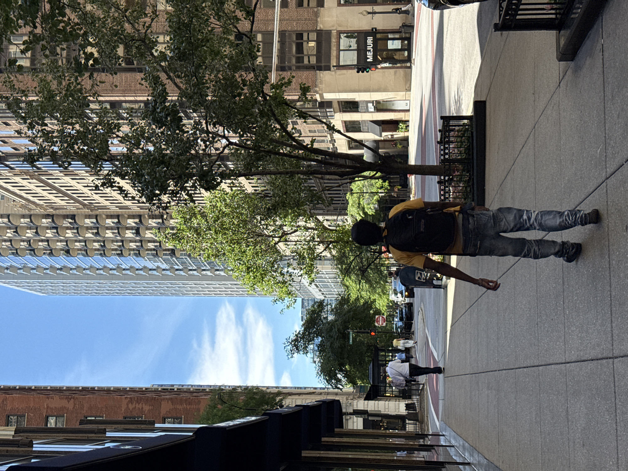 A person wearing a yellow shirt and jeans walks down a sidewalk in Chicago. They have a black backpack on their back. The person is in the foreground of the image, and the background shows buildings and trees. The time of day appears to be daytime, as there is bright sunlight and shadows. The weather seems to be clear, with a blue sky and some white clouds visible.
In the background, there are several tall buildings, some made of brick and others of glass and steel. There are also trees lining the sidewalk, providing shade. A traffic light is visible, indicating that this is a street in a city. The scene appears to be a typical urban environment. The person's posture and gait suggest they are walking casually. No particular emotion is discernible.
There is visible text on a sign in the background that reads "MEJURI". The image is oriented sideways, with the sky at the bottom and the ground at the top.